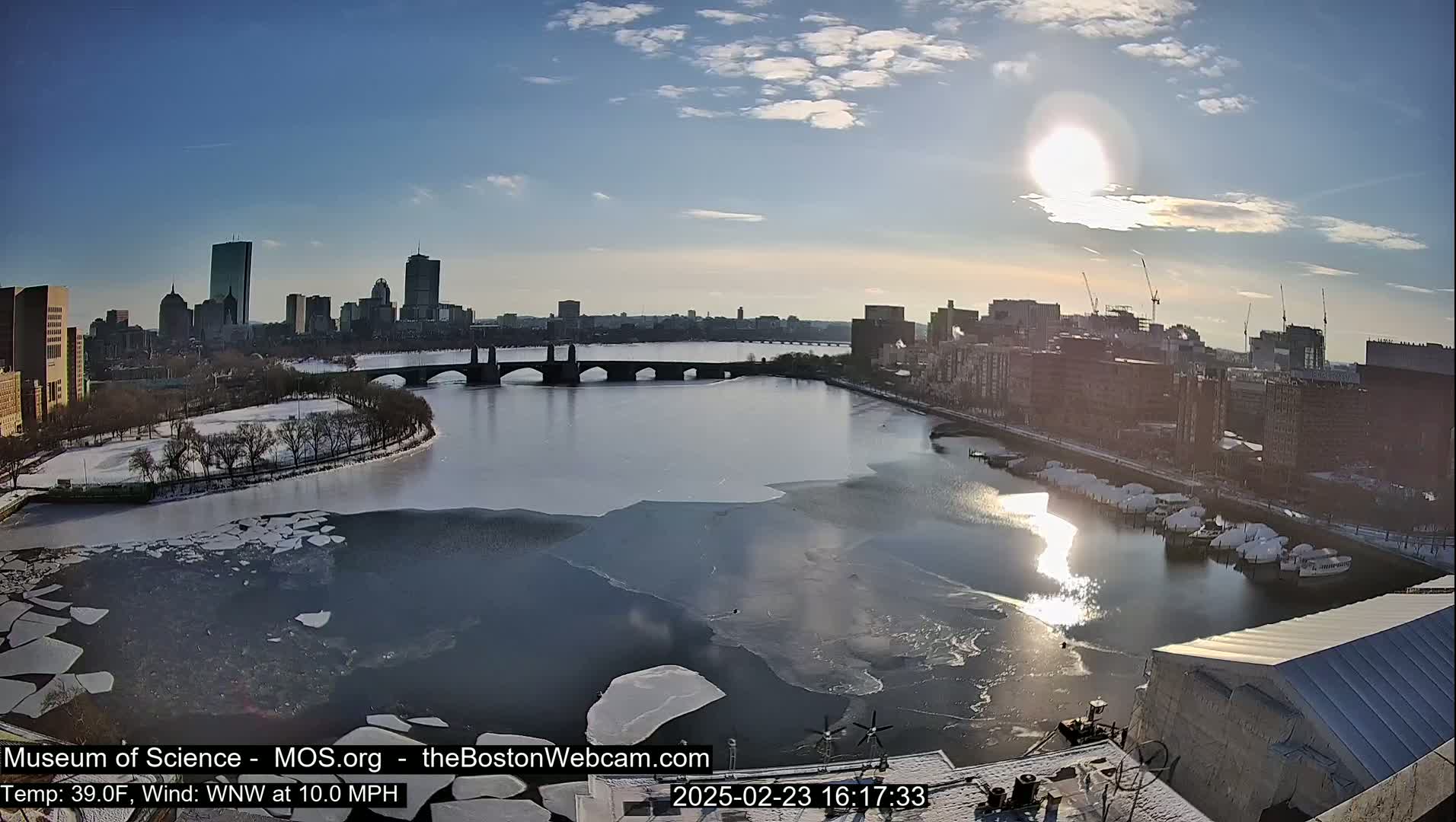 A partially frozen river flows past a city skyline under a bright, sunny sky, with a bridge spanning the river and snow visible on the ground and some boats.