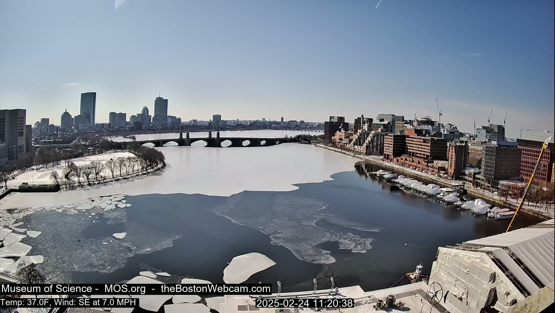 A partially frozen river flows through a city under a clear blue sky, with a bridge spanning the river and snow on the ground.