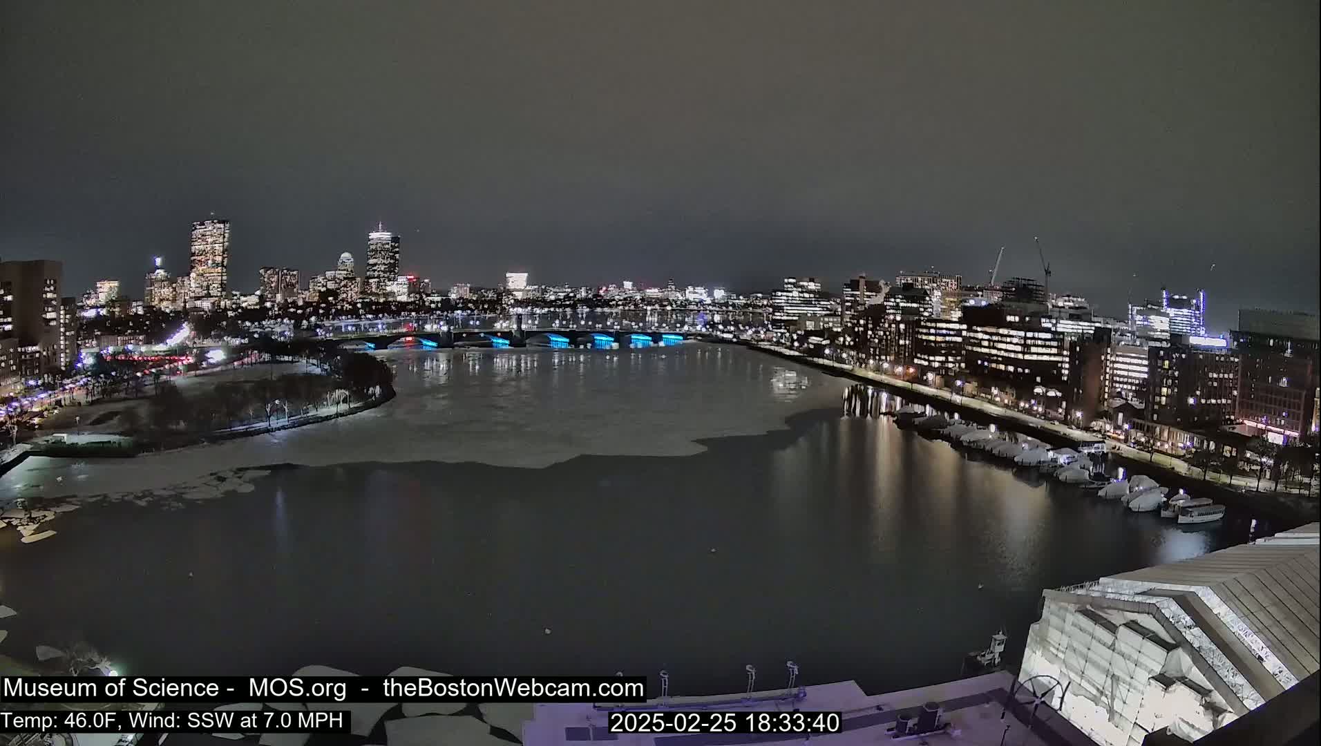 A nighttime cityscape with a partially ice-covered river, illuminated buildings, and boats docked along the shore under a dark sky.