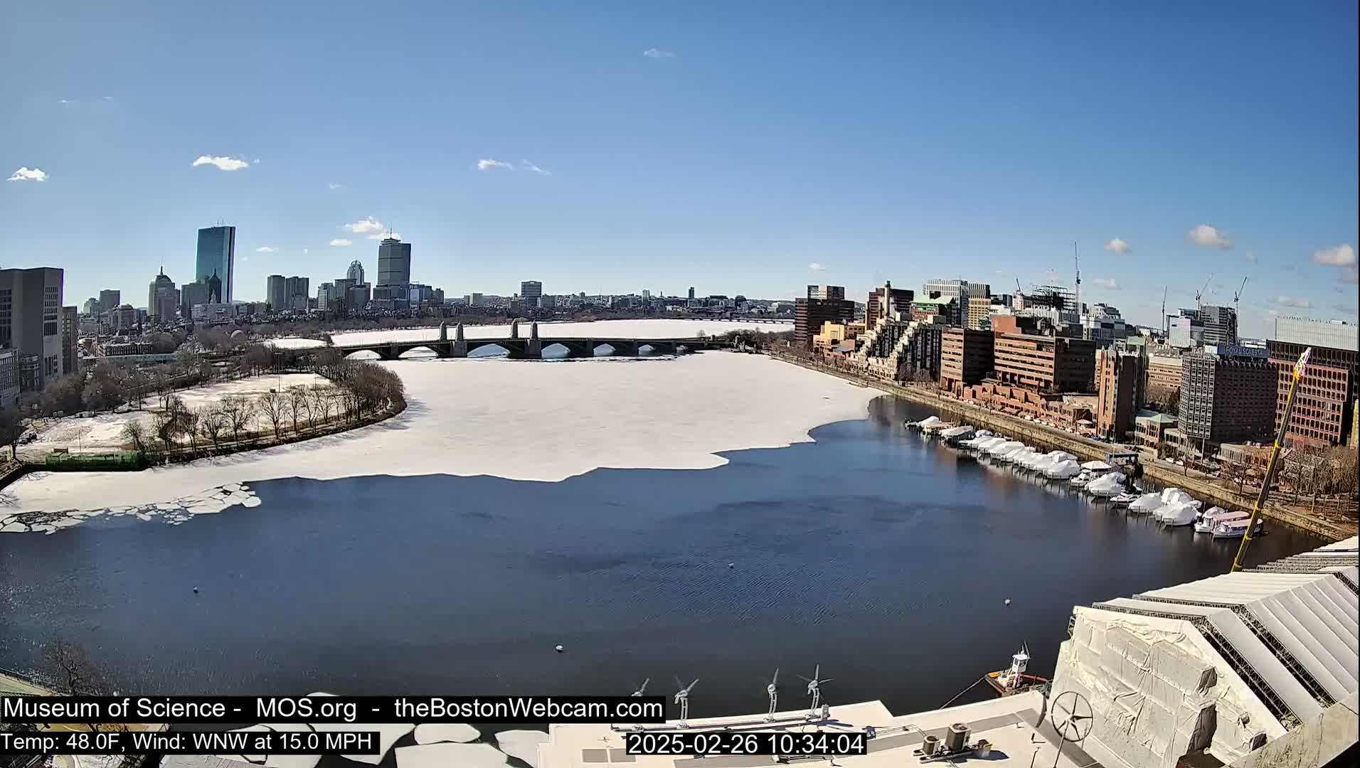 A partly frozen river flows past a city skyline under a clear blue sky, with boats moored along the riverbank.