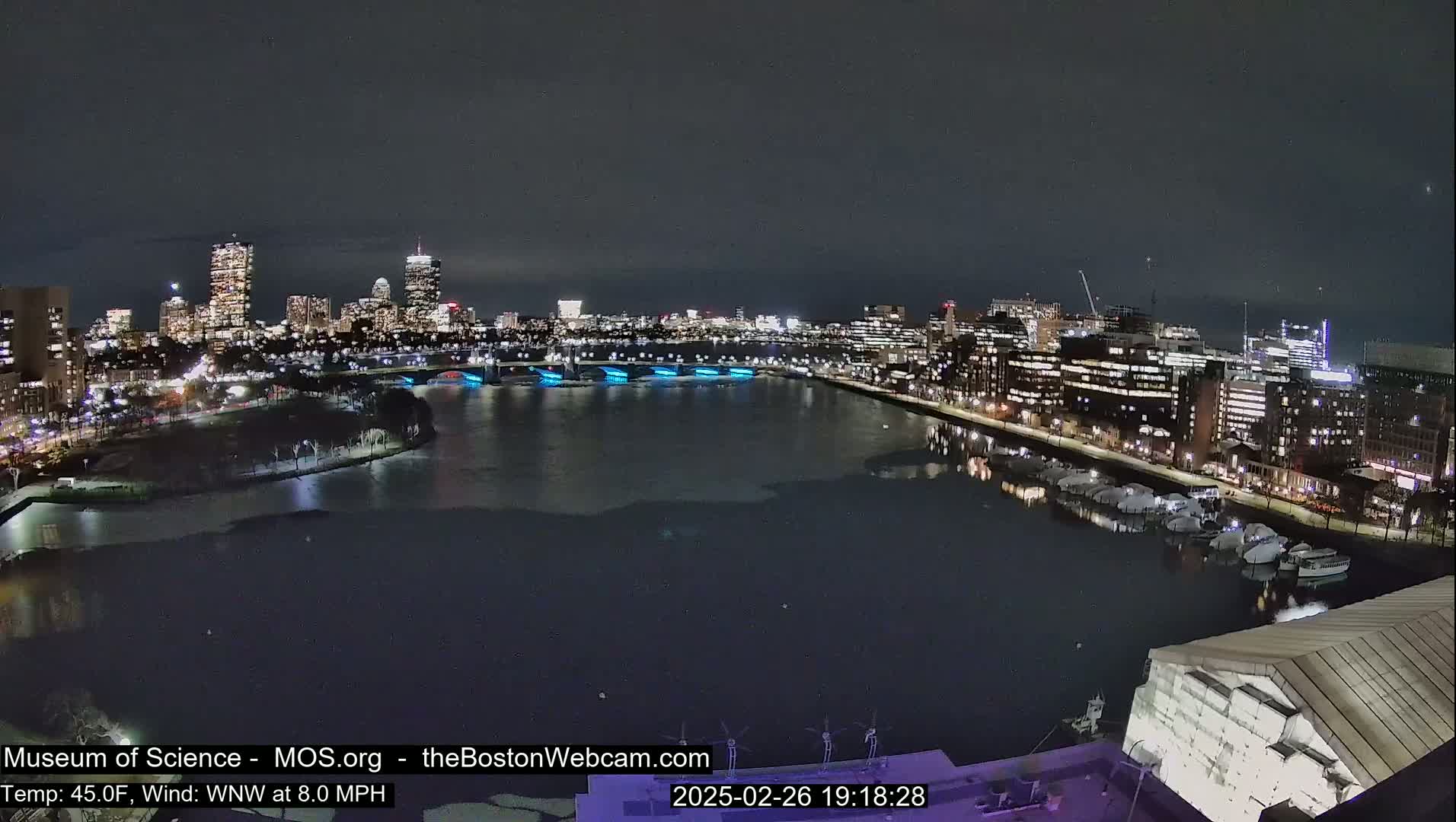 A nighttime cityscape with a partially frozen river reflecting city lights under a clear, dark sky.