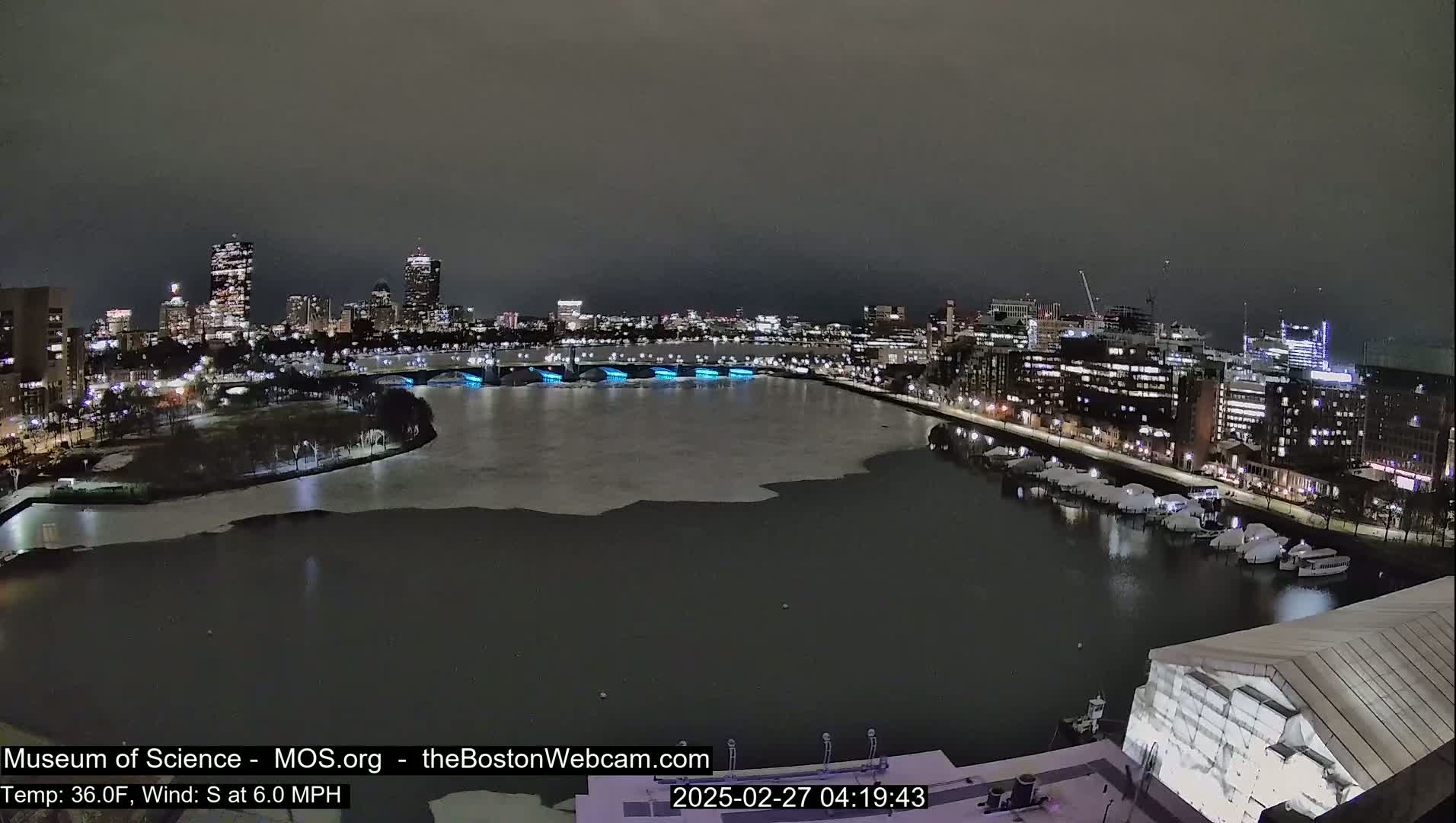 A nighttime view of a partially ice-covered river flowing through a city with buildings illuminated along the banks, under a dark sky.
