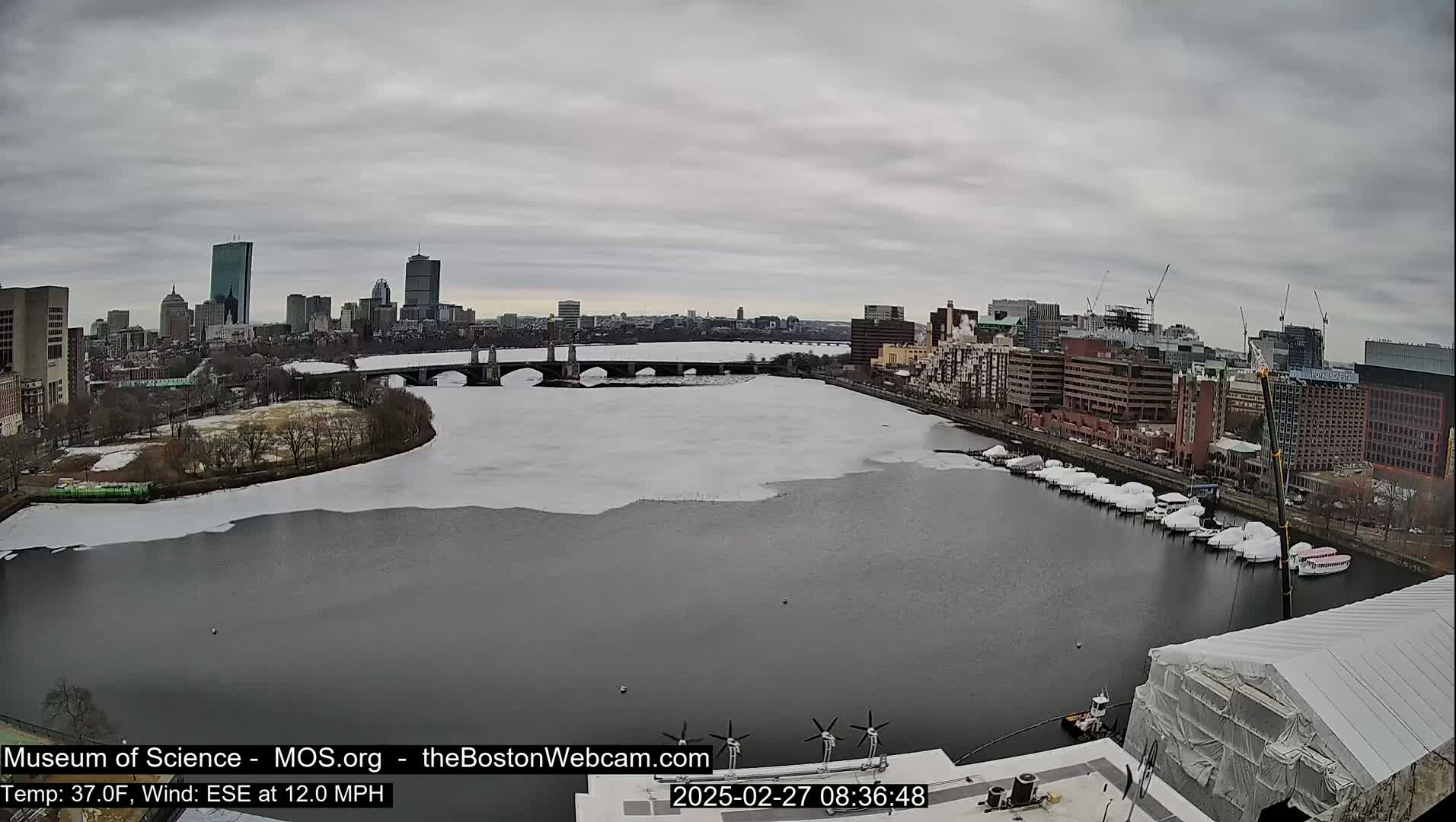 A partially frozen river flows past a city skyline under a cloudy sky, with boats moored along the riverbank and construction visible.