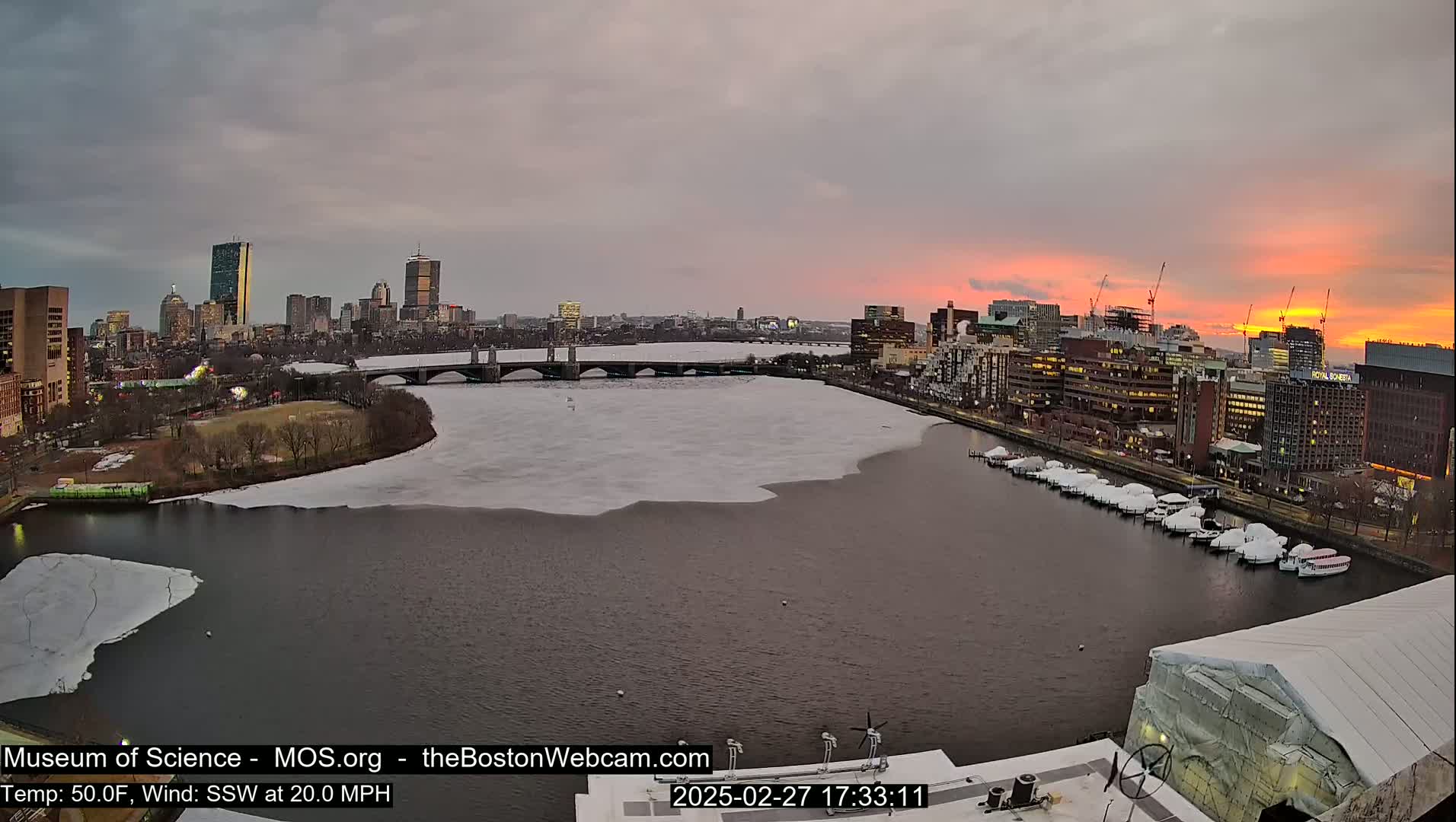 A partially frozen river flows past a city skyline at sunset under a cloudy sky.