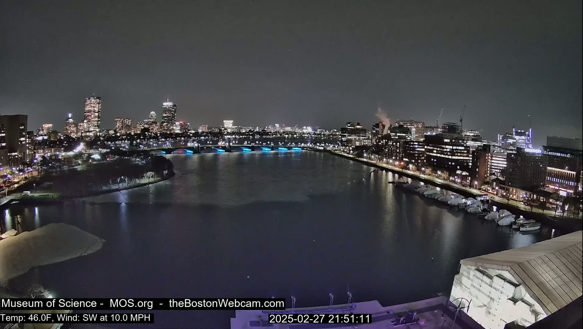 A nighttime view of a city skyline across a partially frozen river, with boats docked along the shore under a dark, clear sky.