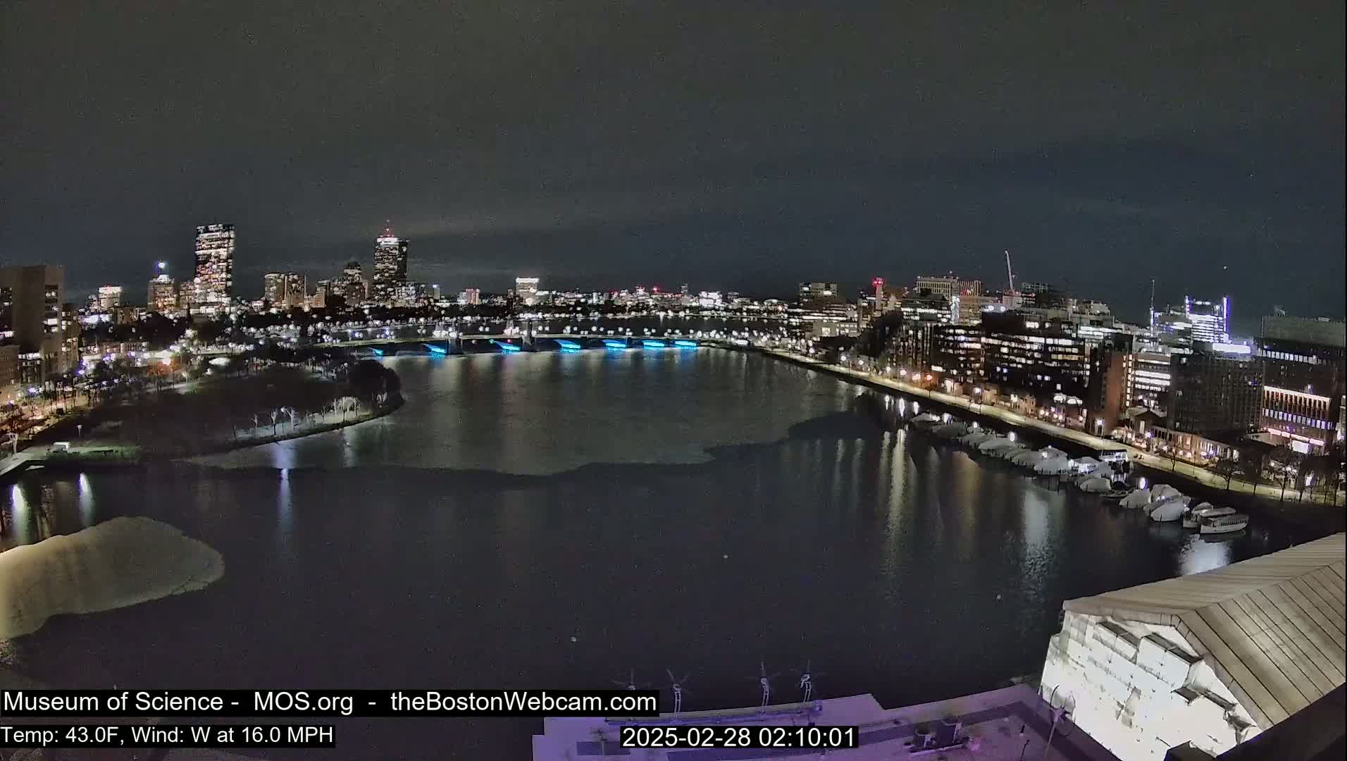 A nighttime cityscape with a partially frozen river, illuminated buildings, and a windy, 43°F temperature.