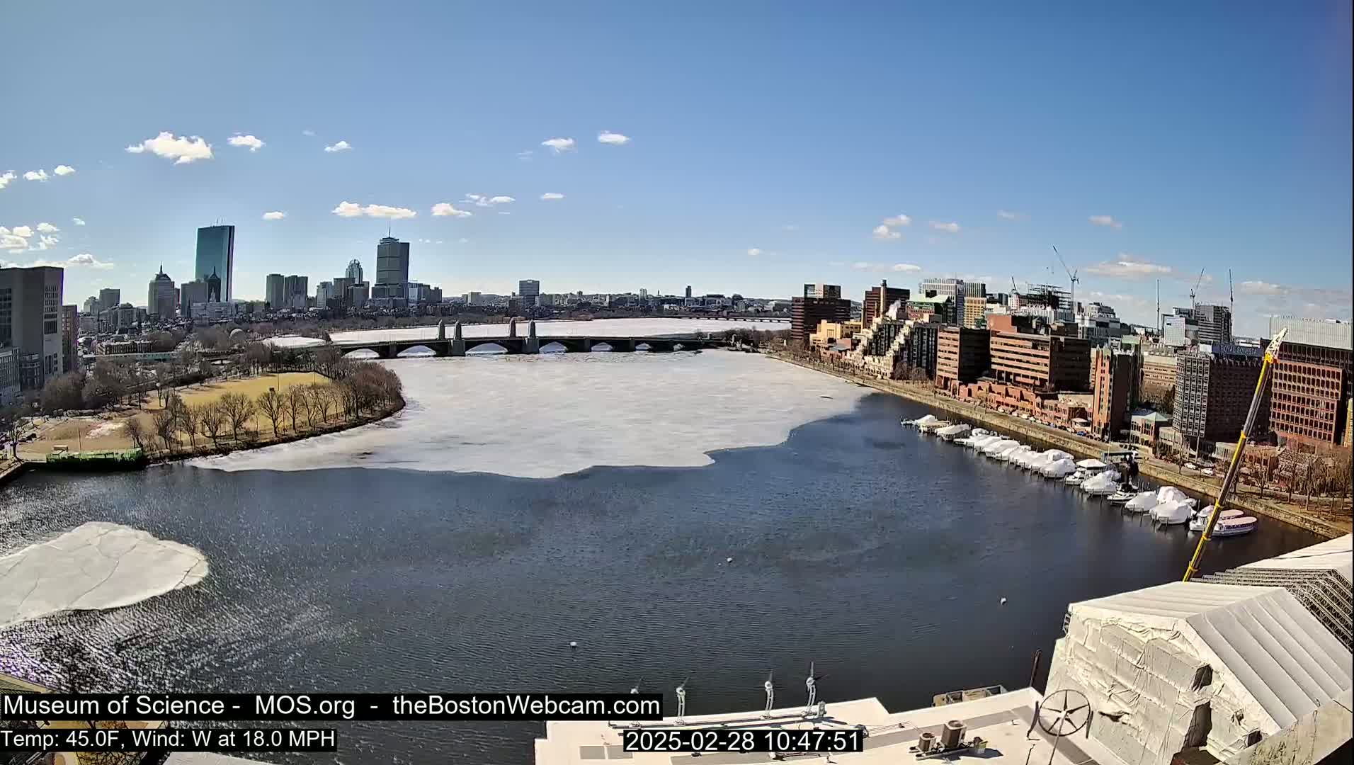 A partially frozen river flows past a city skyline under a clear blue sky, with boats docked along one bank.