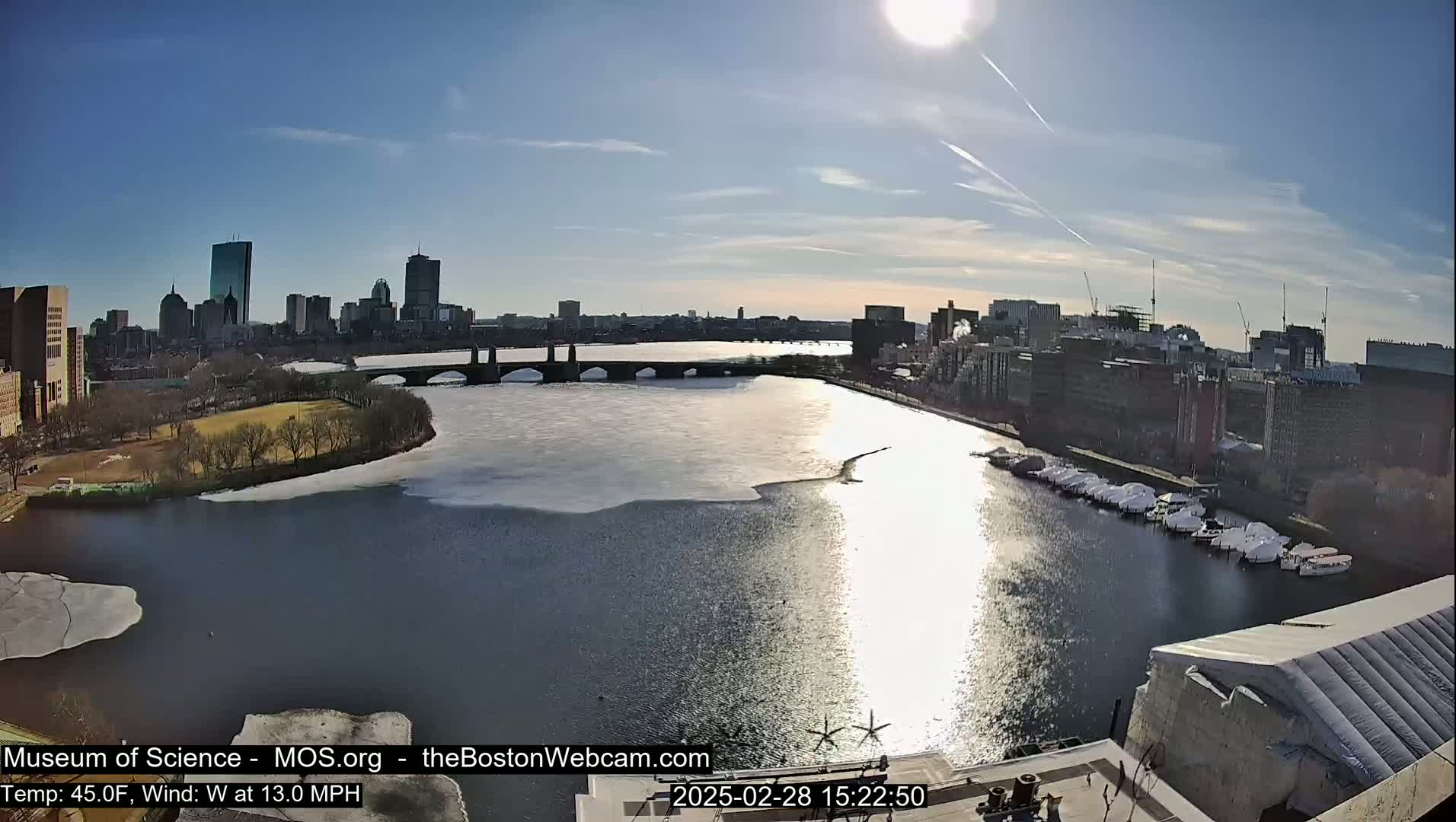 A sunny day with a westerly wind shows a partially frozen river flowing past a city skyline, a bridge, and a marina.