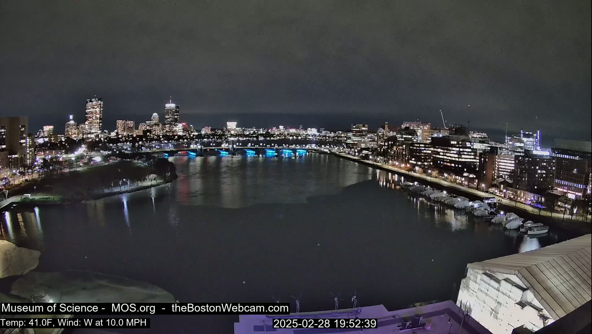 A nighttime view of a city skyline across a dark river with boats docked along the shore, under a cloudy sky.