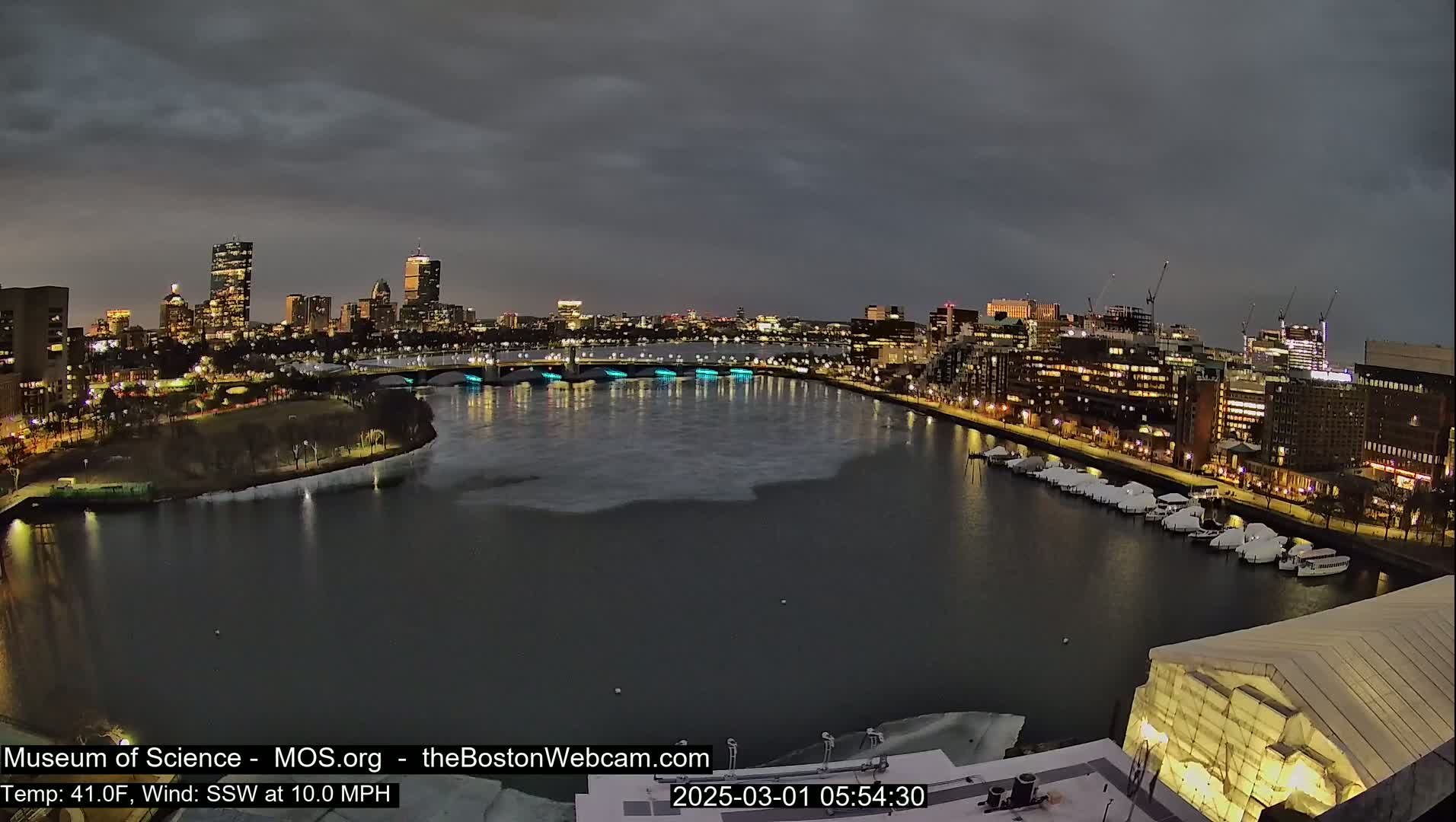 A nighttime cityscape with a partially ice-covered river, under a dark, cloudy sky, showing city lights reflecting on the water and boats moored along the shore.