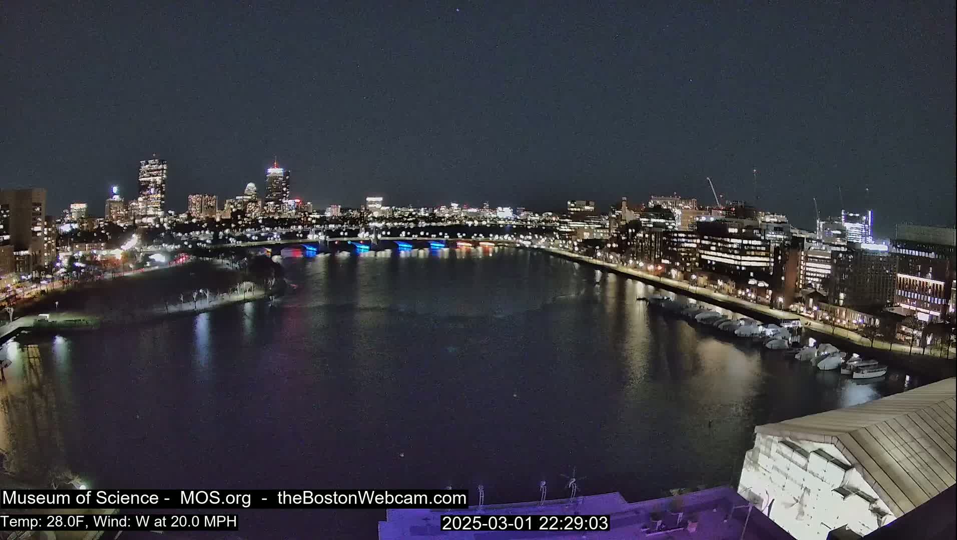 A nighttime view of a city skyline across a dark river, with boats docked along the shore under a clear sky.