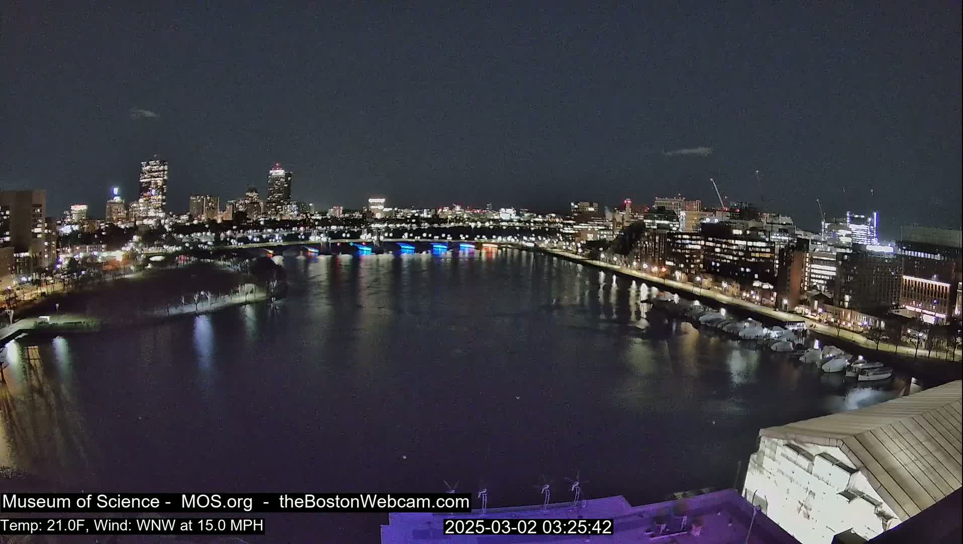 A nighttime view of a city skyline across a body of water, with boats docked along the shore under a clear, dark sky.