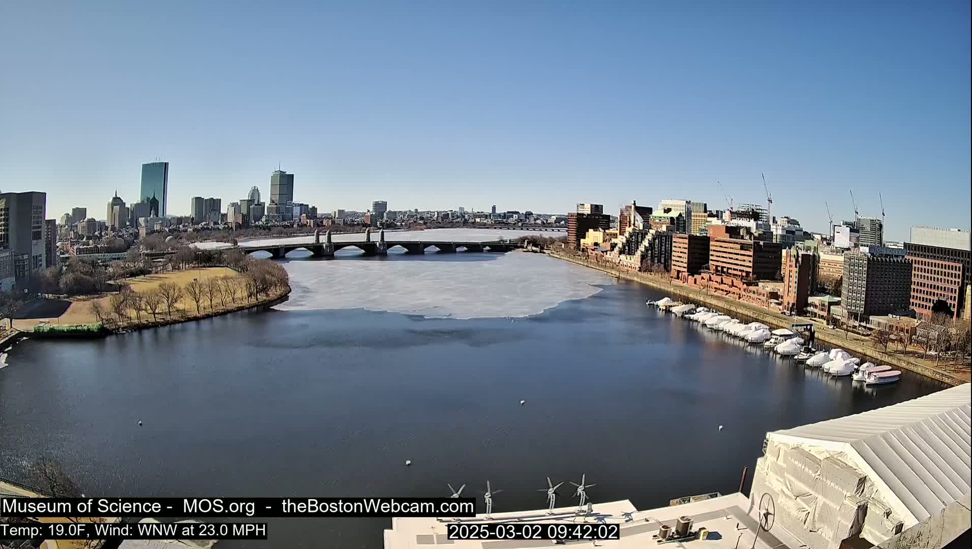 A partly icy river flows beneath a bridge, past a city skyline under a clear, sunny sky.