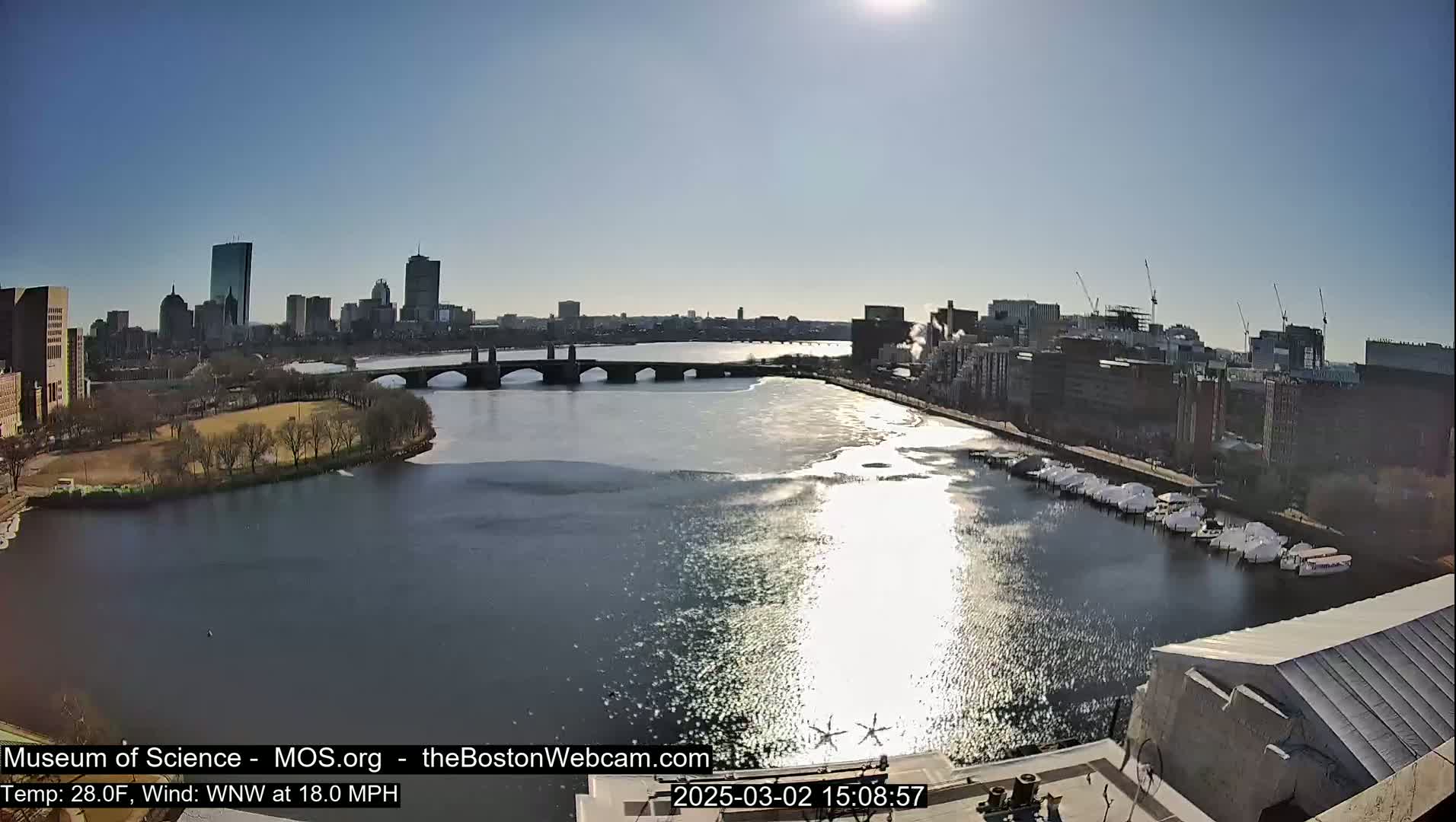 A sunny day reveals a cityscape across a partly ice-covered river, with a bridge and docked boats in the foreground.