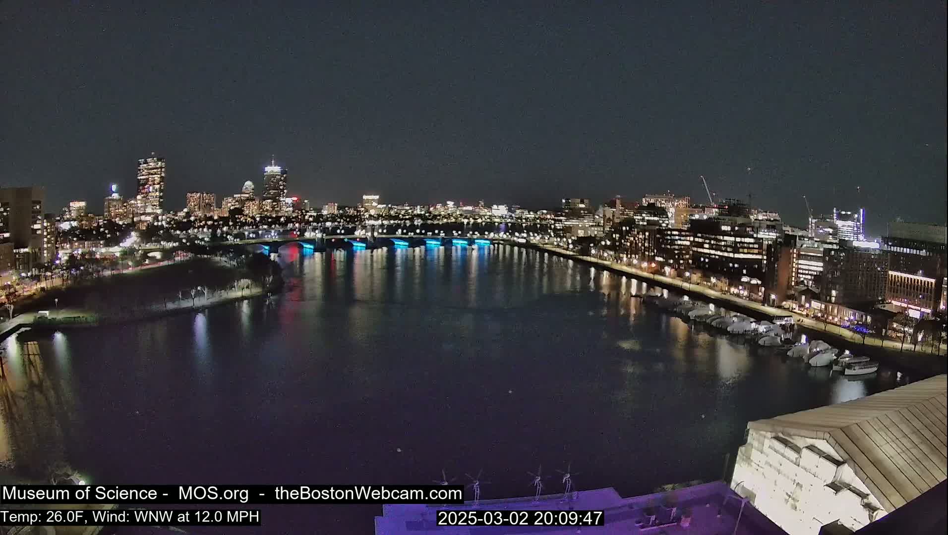 A nighttime view of a city skyline across a dark river, with boats docked along the waterfront under a clear sky, and a 26°F temperature with a 12 mph west-northwest wind.