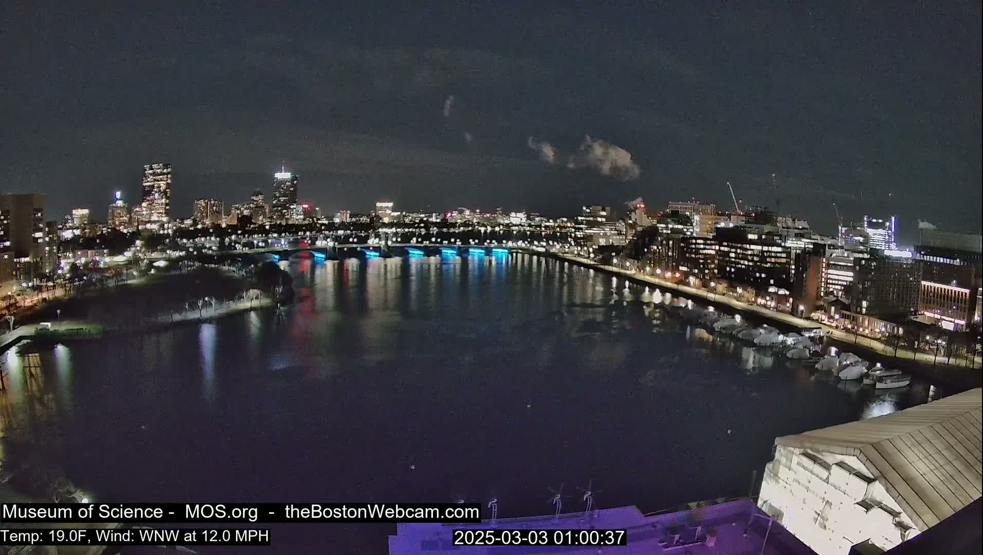 A nighttime view of a city skyline across a river, with a bridge illuminated in blue, under a partly cloudy sky, and a wind of 12 mph.