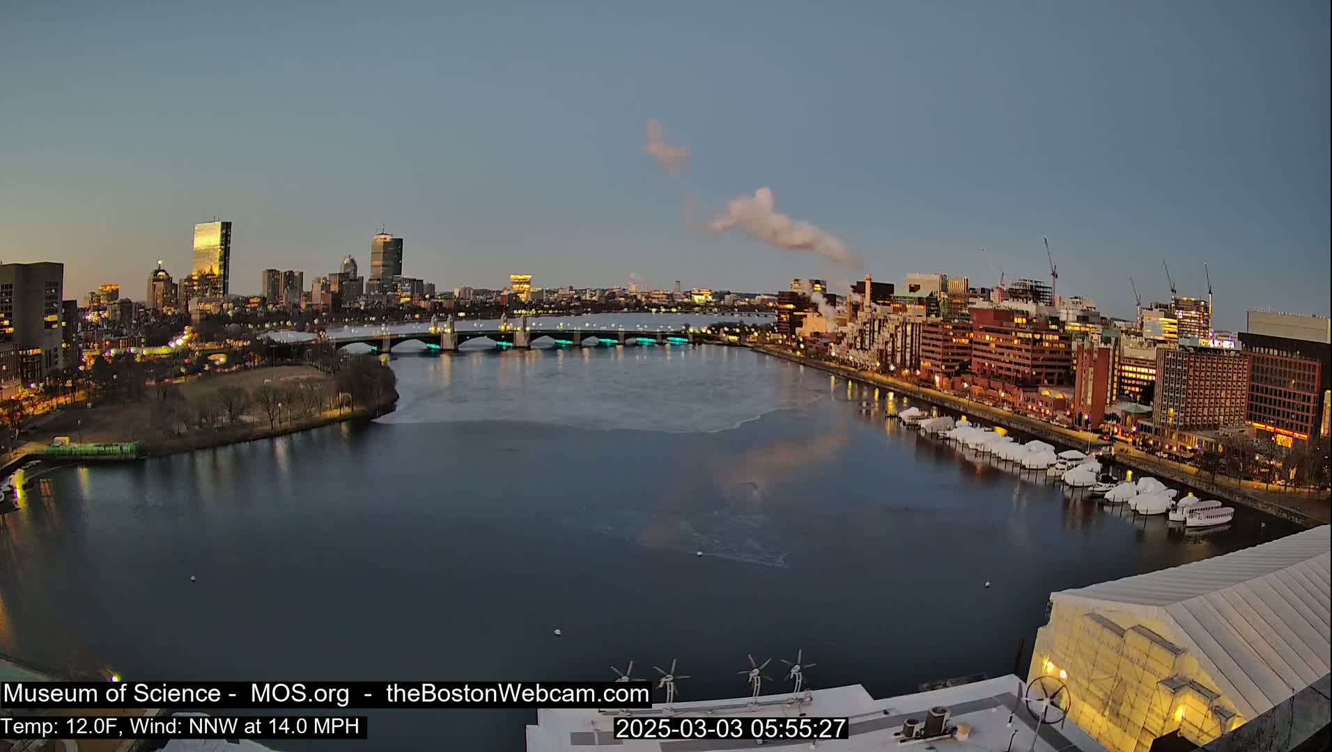 A city skyline at twilight is partially visible across a partially frozen river, with boats docked along the shore and a structure with a large white canopy in the foreground under a clear, cool sky.