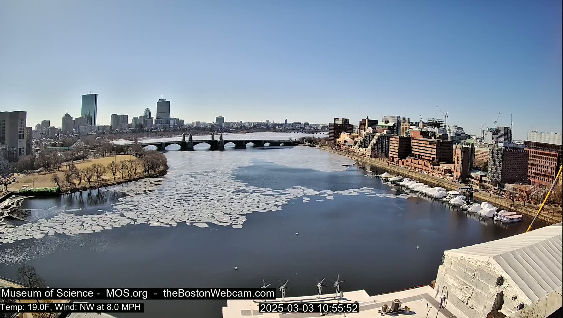 A partially ice-covered river flows past a city skyline under a clear blue sky, with boats docked along the riverbank.