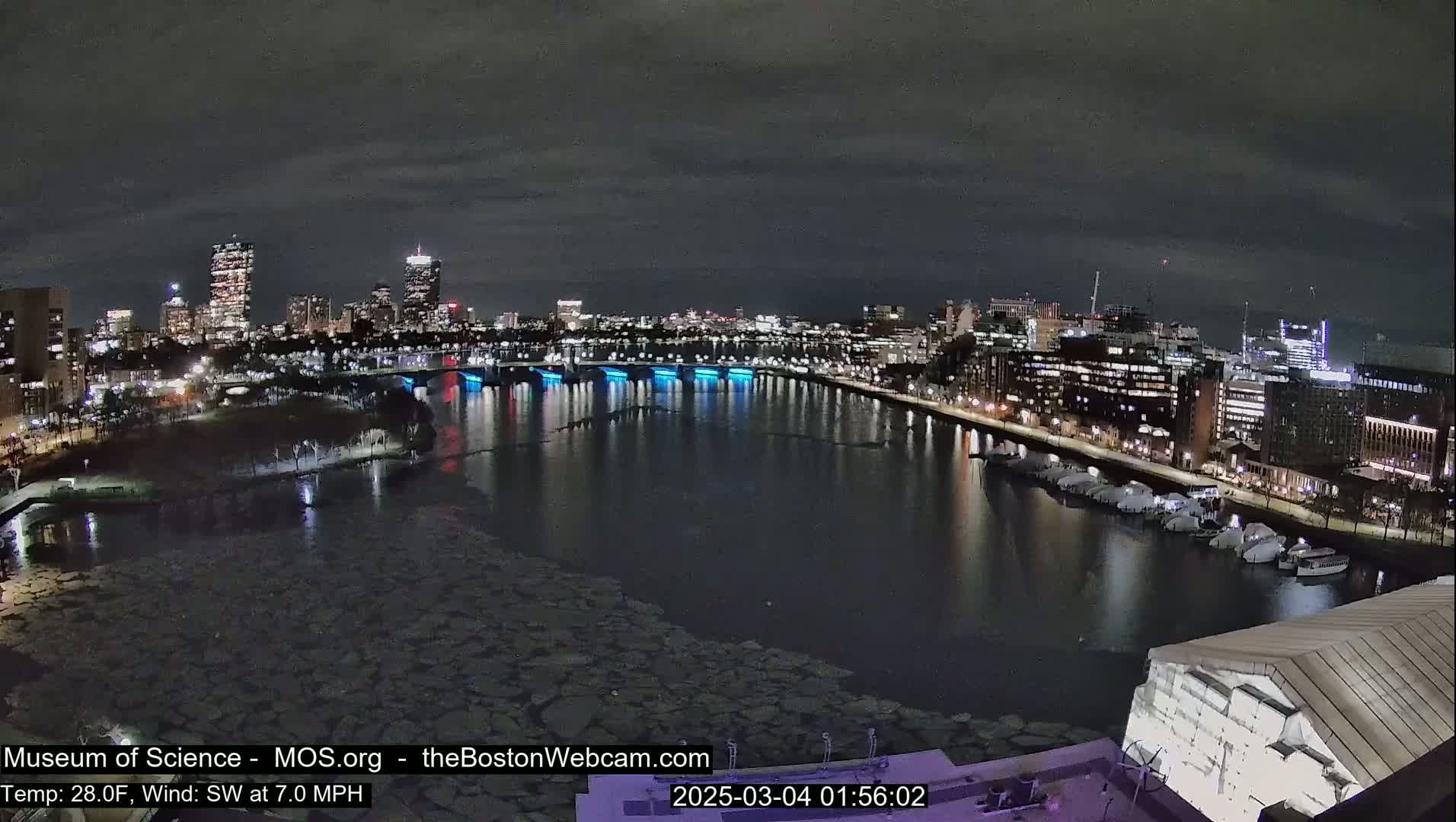 A nighttime cityscape with a river partially frozen over, lit by city lights under a dark, cloudy sky.