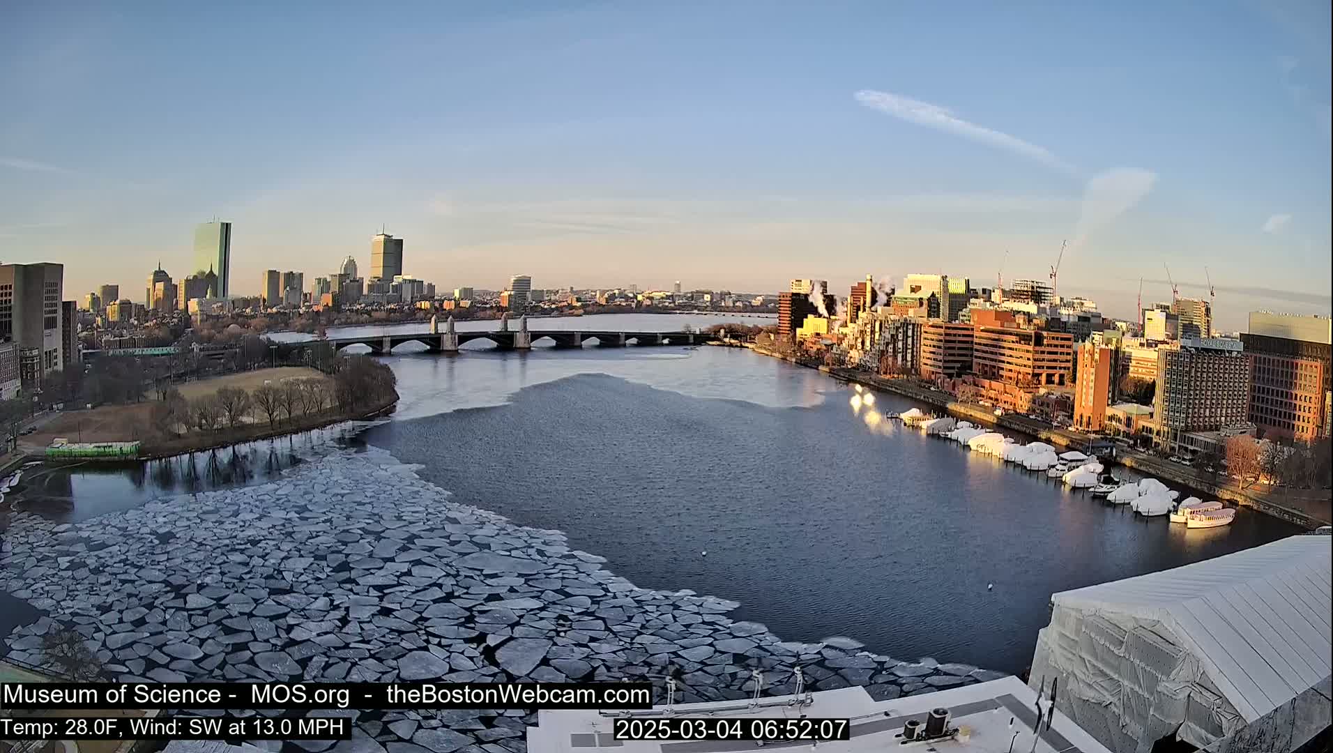 A partially frozen river flows past a city skyline under a clear, bright sky.