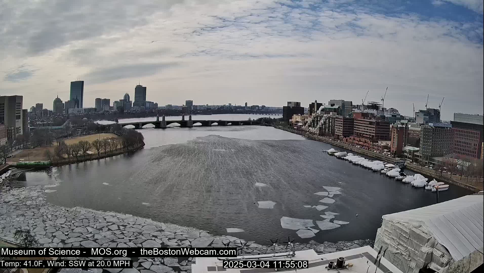 A city skyline is visible across a river partially covered with broken ice floes under a partly cloudy sky.