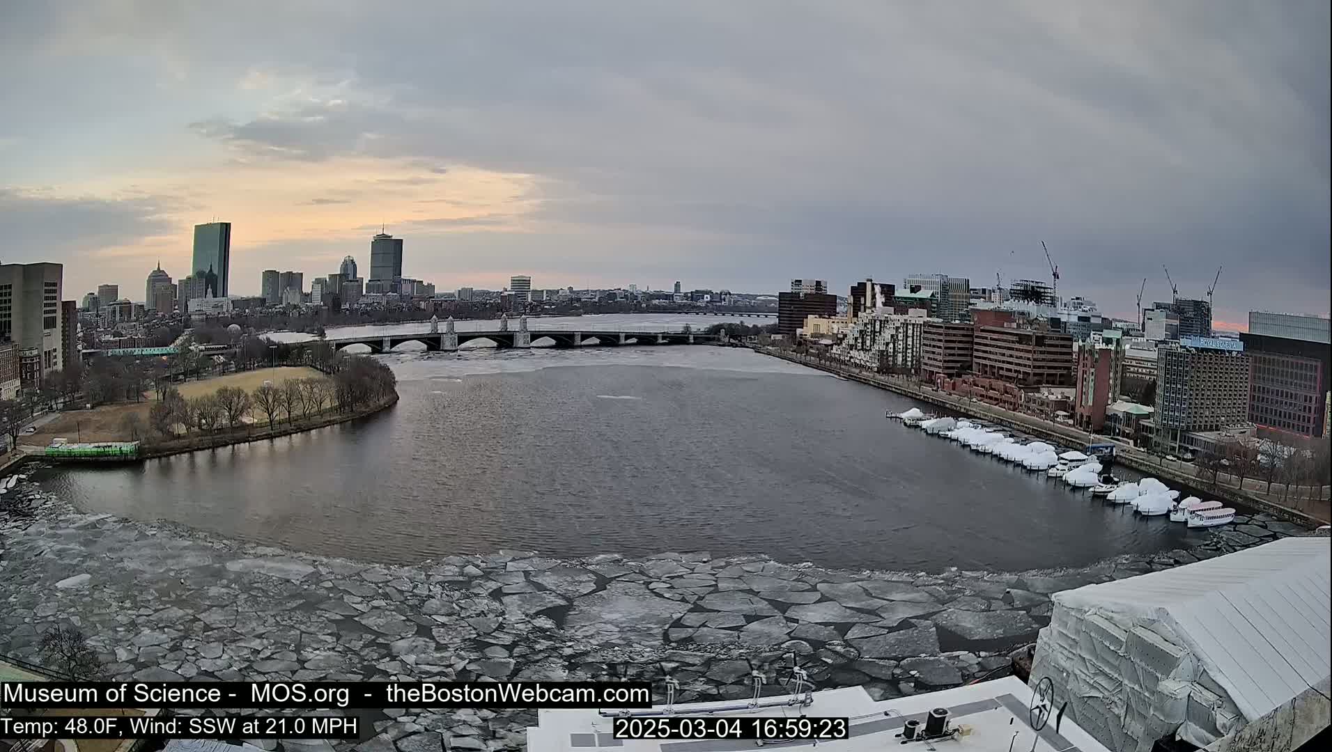 A city skyline is visible across a river partially covered in ice floes, under an overcast sky.