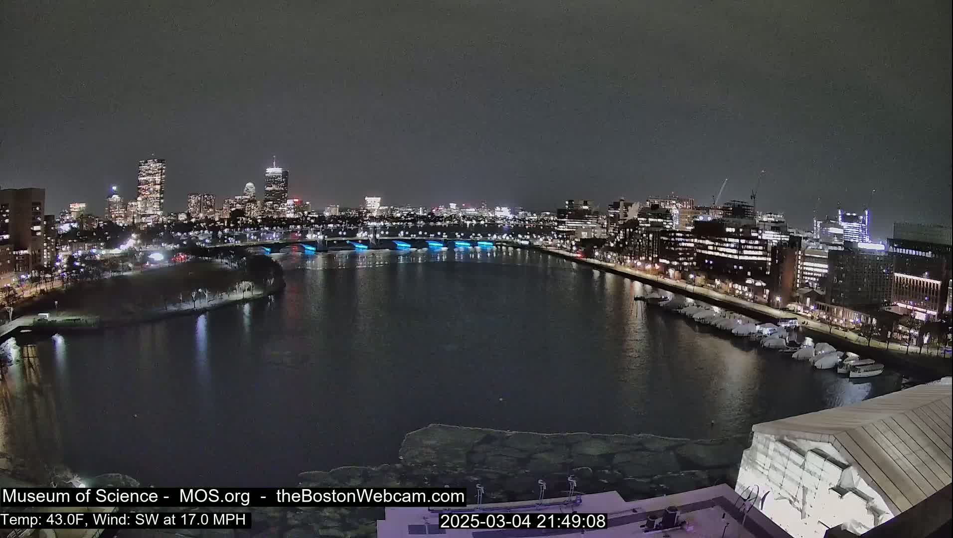 A nighttime view of a city skyline across a river partially covered in ice, under a dark sky with a southwest wind at 17 mph.