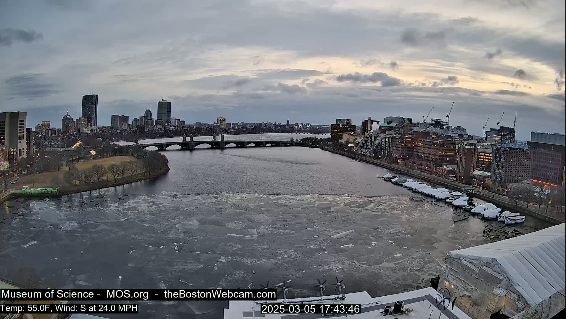 A partially frozen river flows past a city skyline under a cloudy, twilight sky.