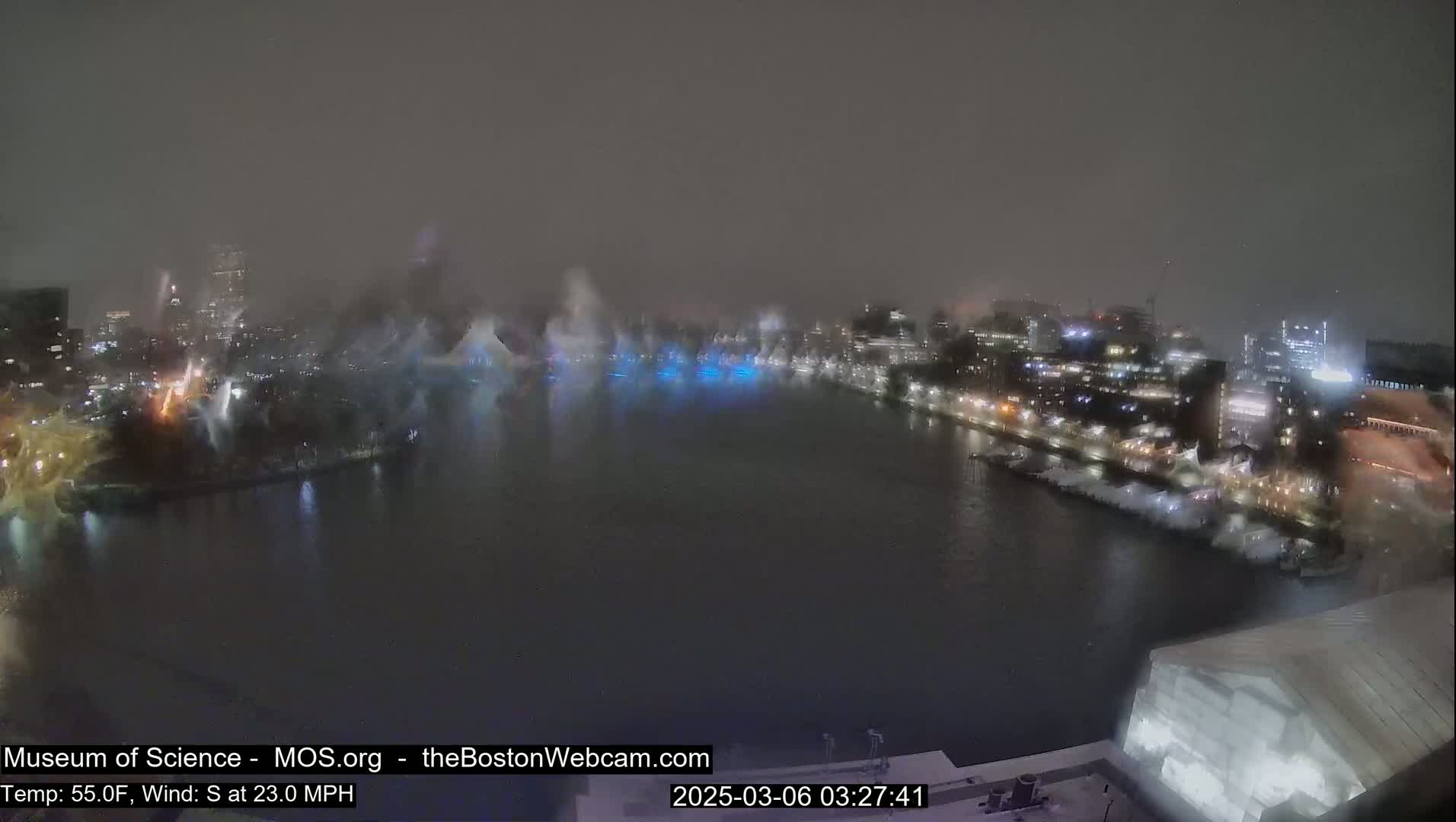 A nighttime, snowy scene shows a waterfront city with lights and what appears to be a water show or fireworks display over a dark body of water, under a hazy sky with a 23 mph south wind.