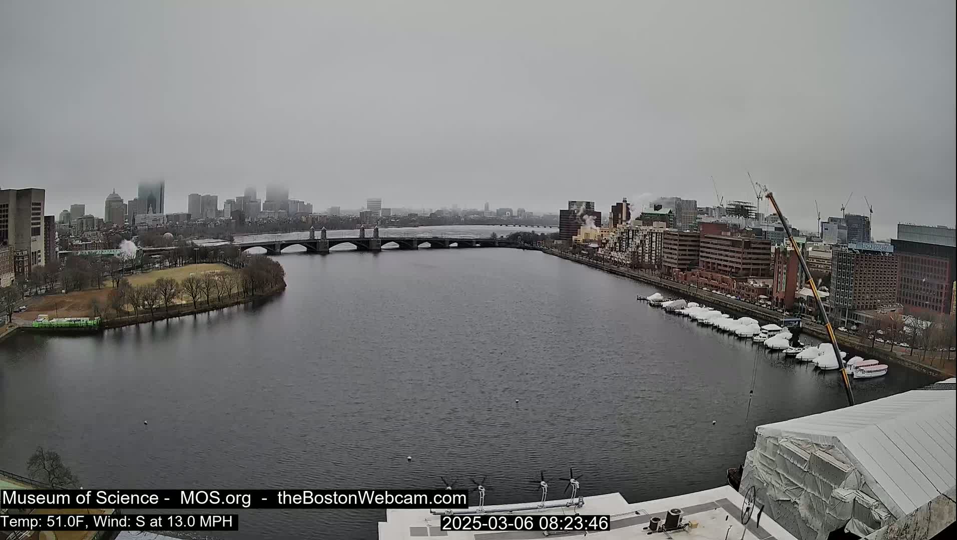 A foggy view of a city skyline across a river with a bridge, boats docked along the shore, and a partially covered building in the foreground.
