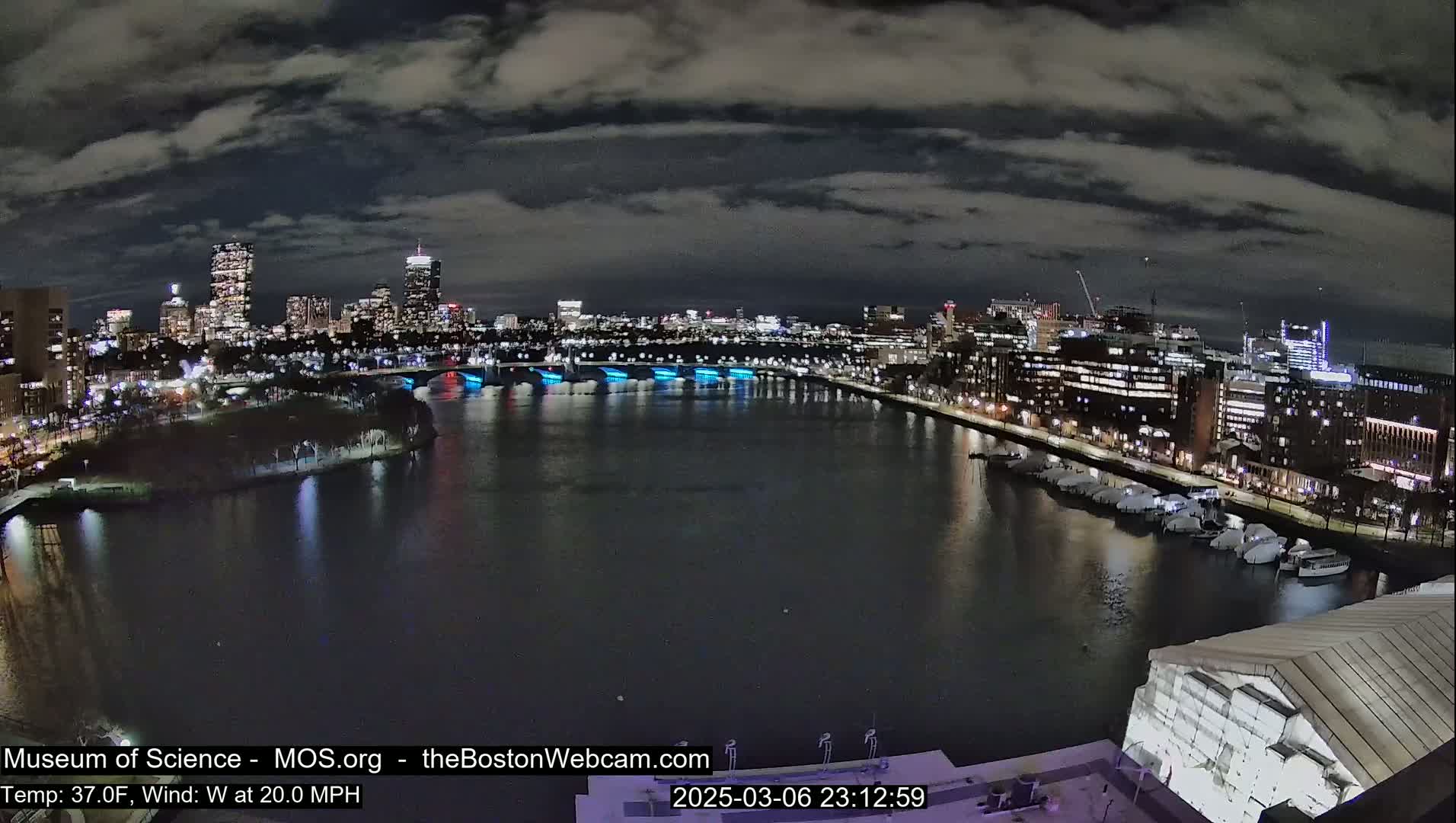 A nighttime cityscape with a river reflecting city lights under a cloudy sky.