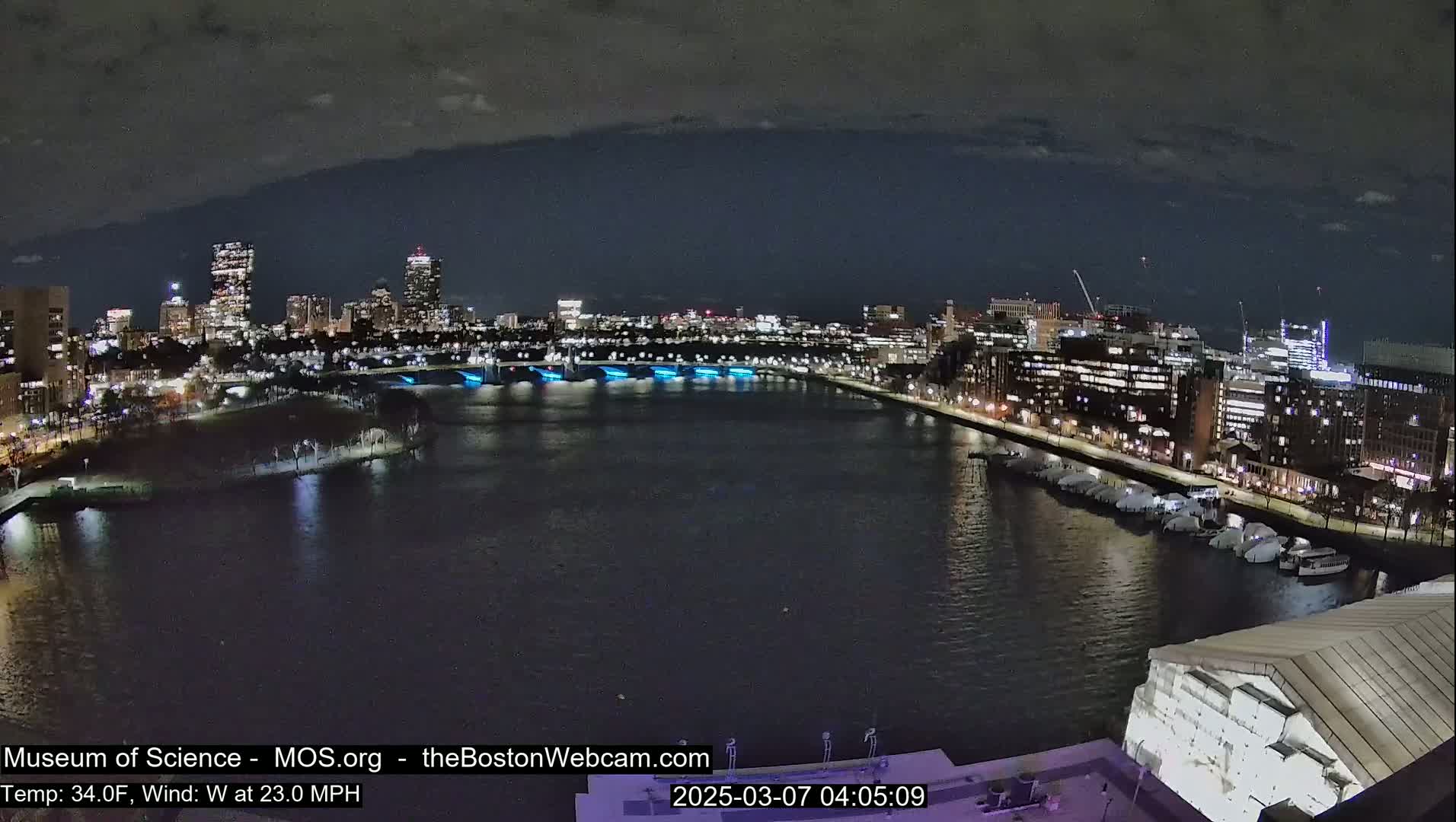 A nighttime cityscape with a body of water, lit city buildings, and a bridge with blue lights along its side, under a dark, cloudy sky.