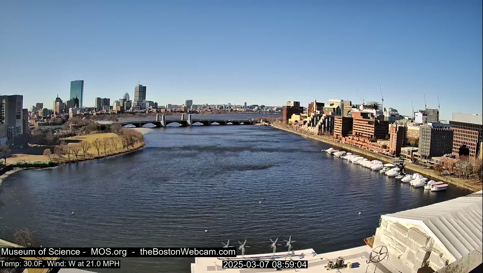 A clear, sunny day shows a cityscape across a wide river with a bridge, boats docked along the shore, and some wind turbines in the foreground.