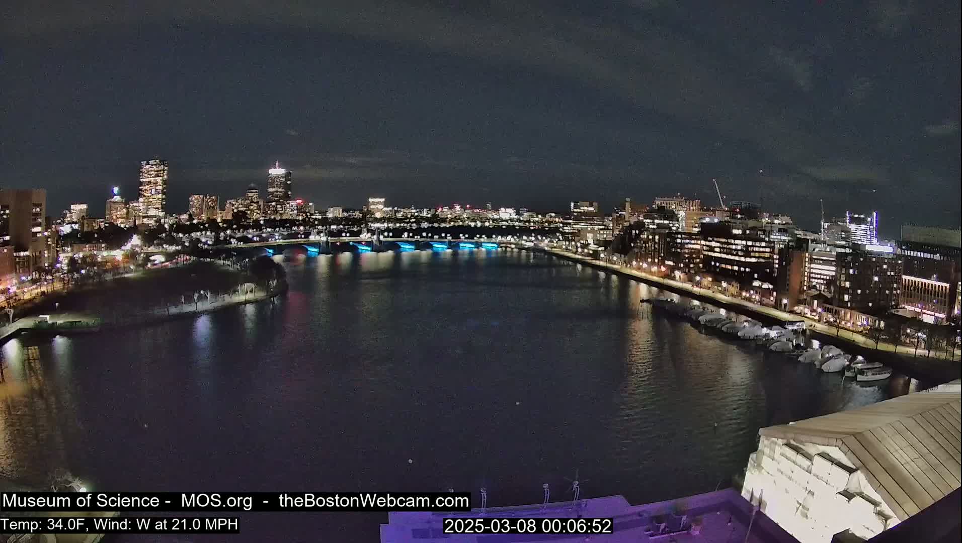 A nighttime, high-angle view of a city waterfront, with a river reflecting the city lights under a dark sky and a moderate west wind.