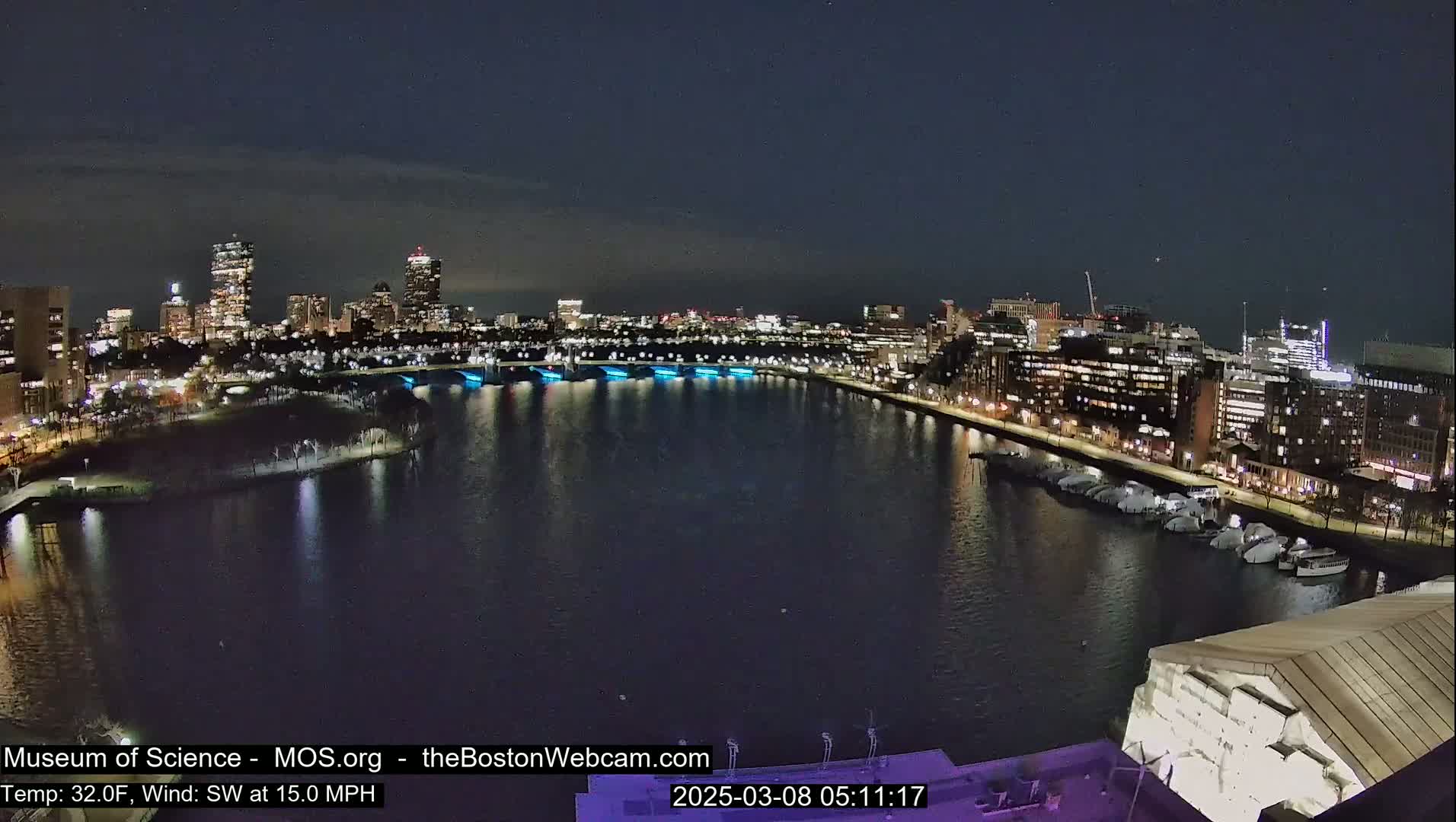A nighttime view of a city skyline across a dark river, with a 32°F temperature and a 15 mph southwest wind.