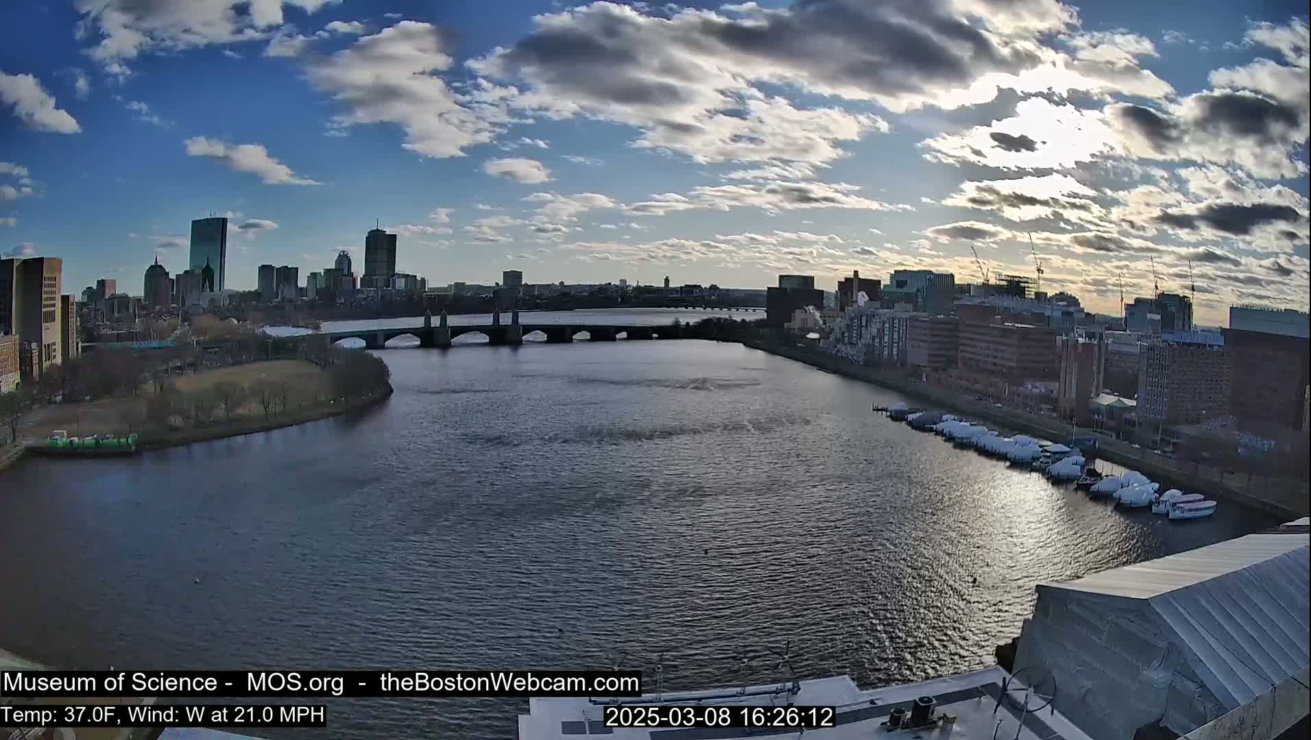 A partly cloudy day shows a city skyline across a river with a bridge and boats docked along the shore.