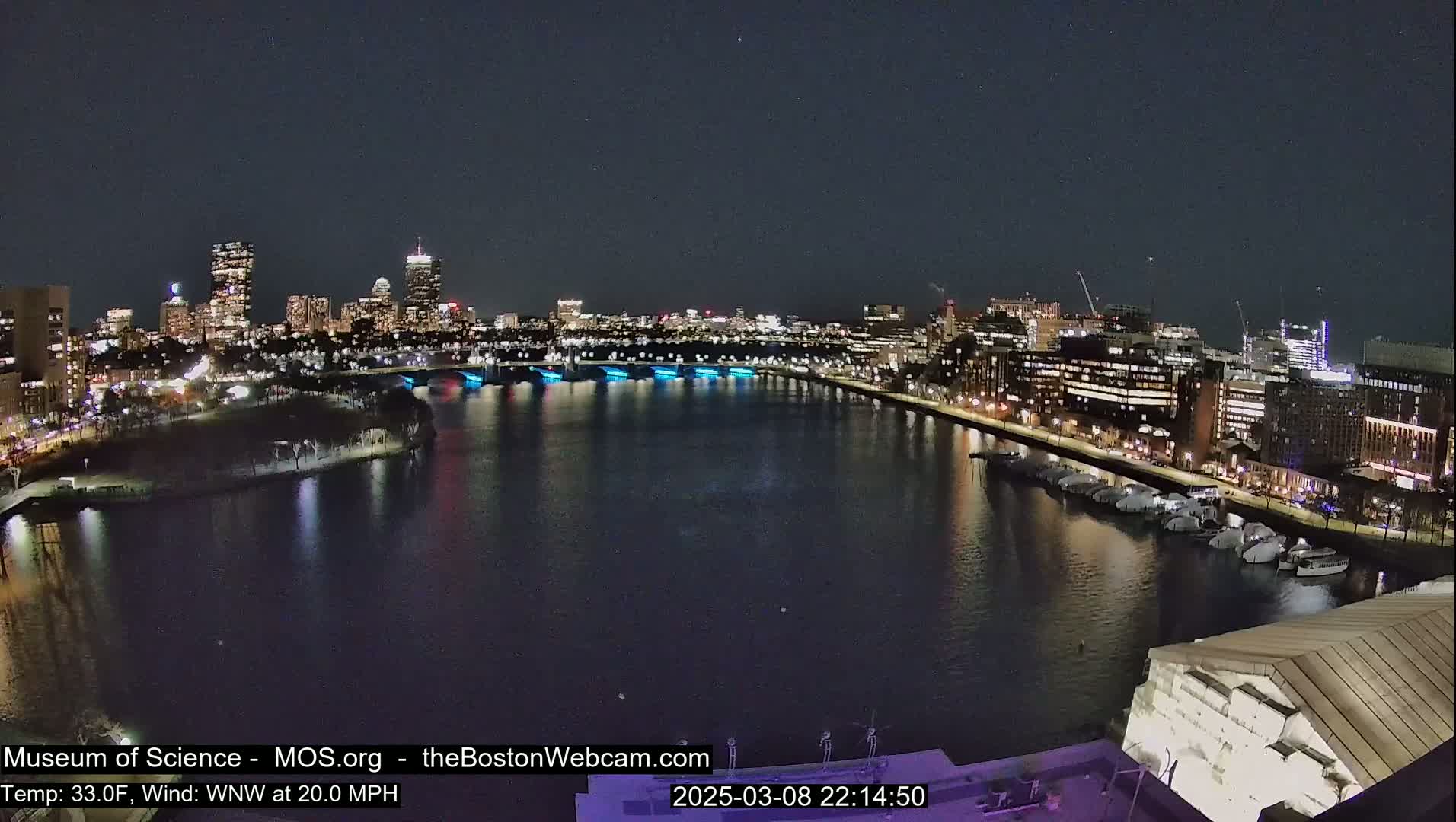 A nighttime cityscape with a body of water reflecting city lights, under a clear, dark sky; a moderate wind is present.