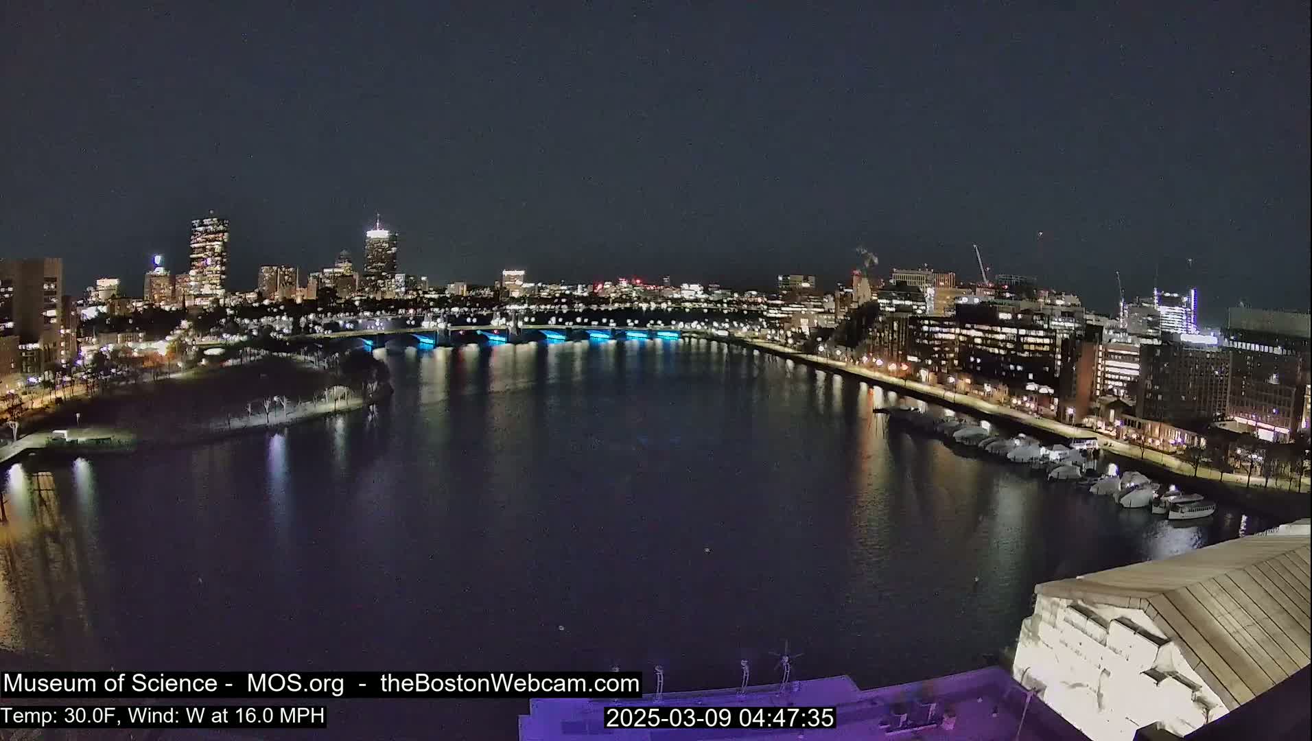 A nighttime view of a city skyline across a dark river, with a bridge illuminated in blue, under a clear sky and a 30°F temperature with a 16 mph west wind.