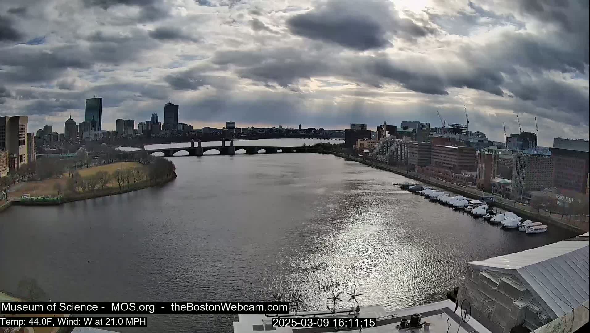 A city skyline with numerous buildings is visible across a wide river under a cloudy sky, with a bridge connecting the city to a waterfront area where boats are docked.