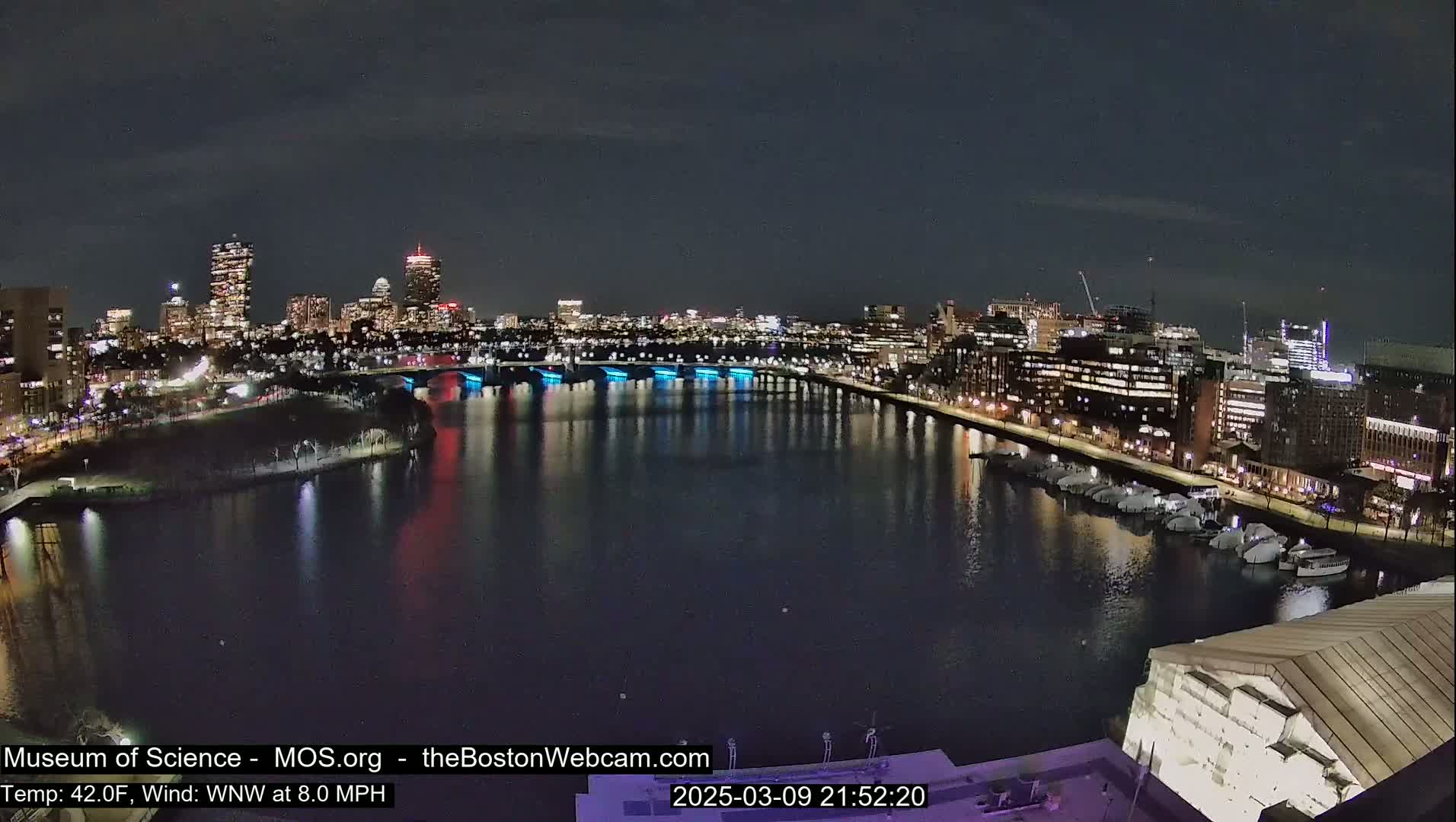 A nighttime view of a city skyline across a body of water, with boats moored along the shore under a clear, dark sky.