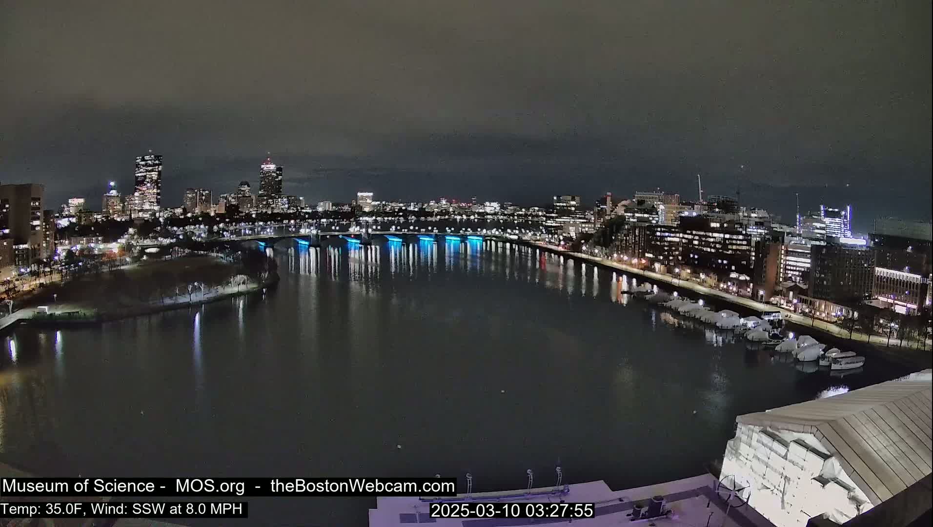 A nighttime view of a city skyline across a dark river, with city lights reflecting on the water under a dark, overcast sky.