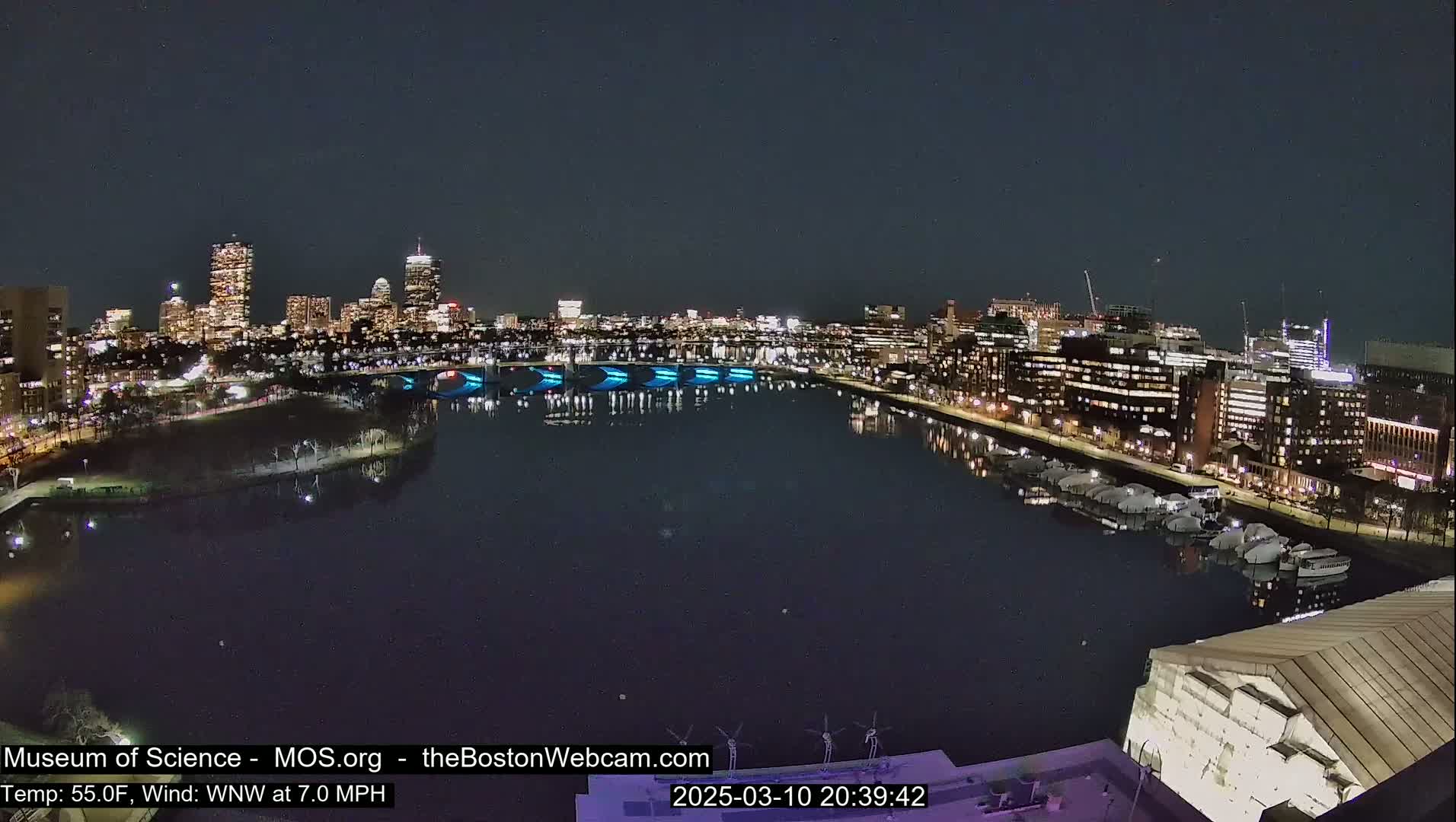 A nighttime view of a city skyline across a calm body of water, with a bridge illuminated in blue.