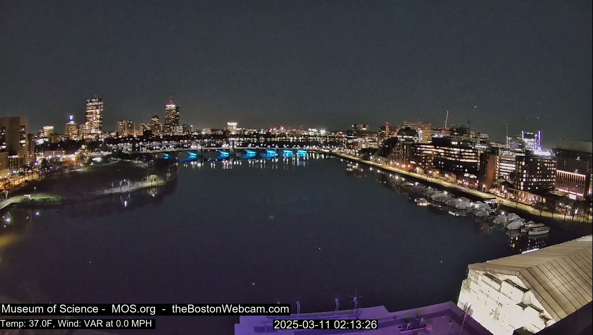 A nighttime view of a city skyline across a dark river, with a bridge illuminated in blue.