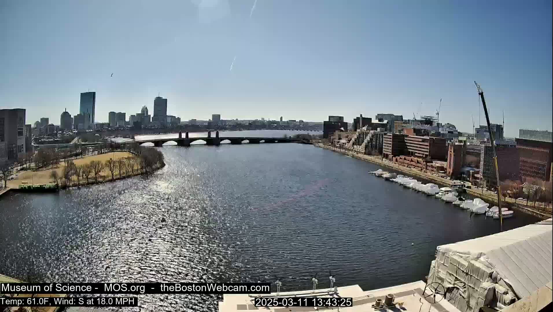 A sunny day shows a cityscape across a river with a bridge, boats docked along the shore, and a construction crane.