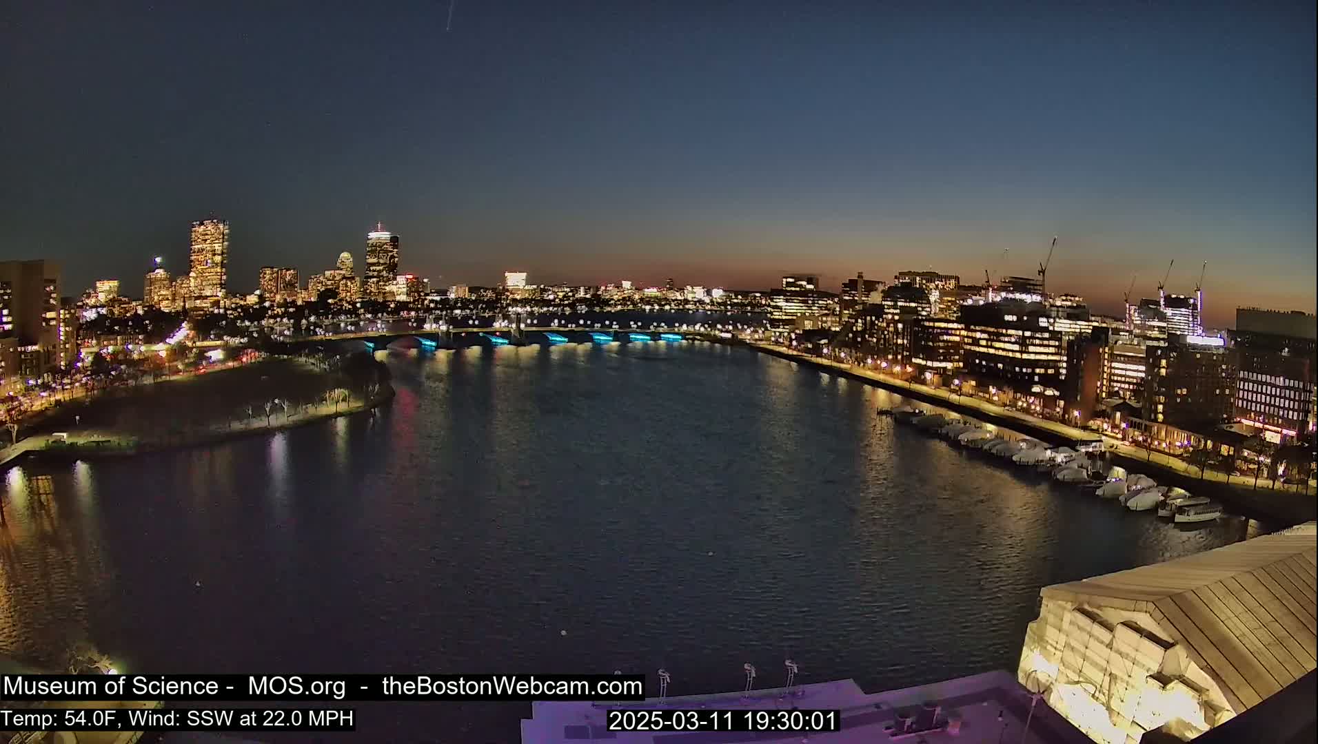A nighttime view of a city skyline across a calm body of water, with boats docked along the shore under a clear, dark sky.