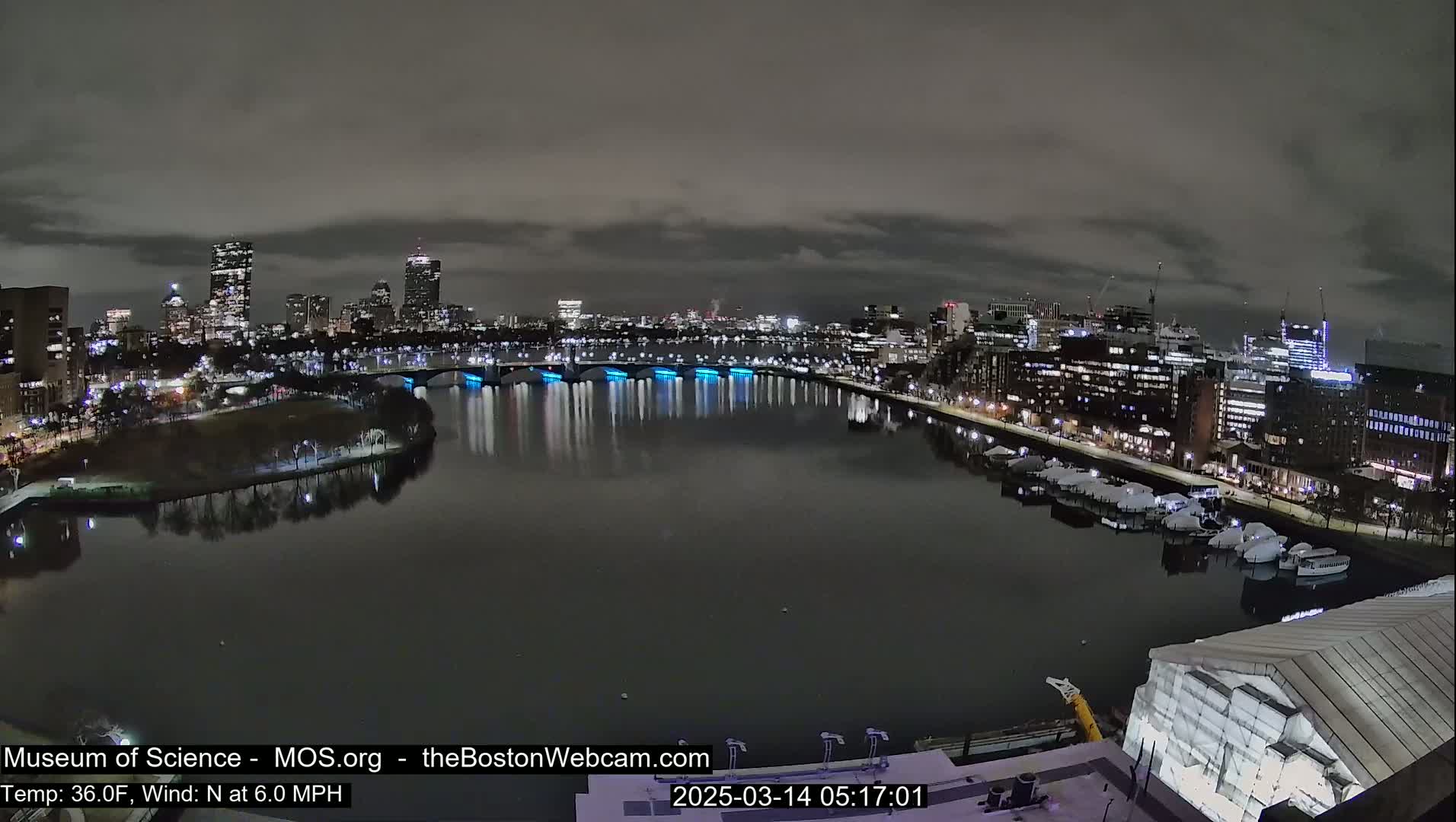 A nighttime view of a city skyline across a calm river, under a cloudy sky.