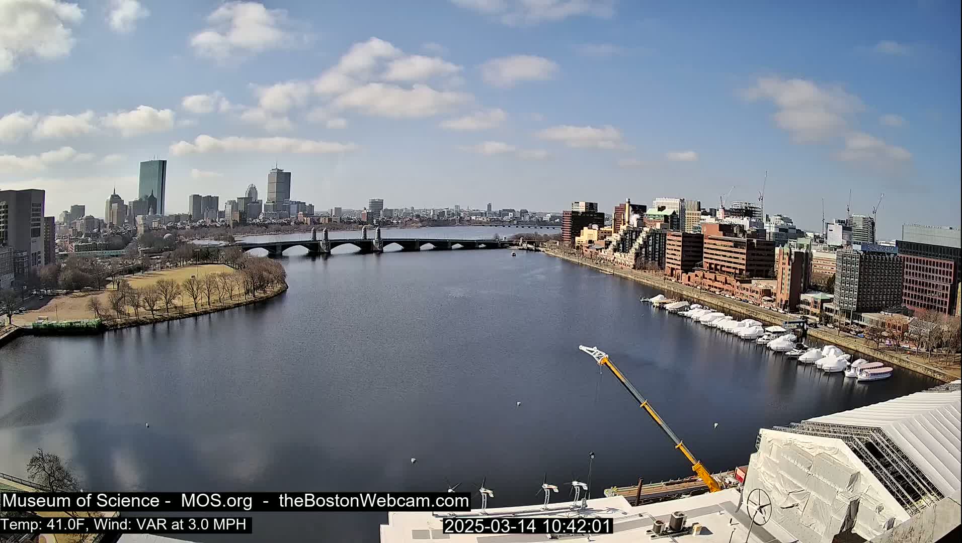 A city skyline with a river, bridge, and docked boats under a partly cloudy sky.