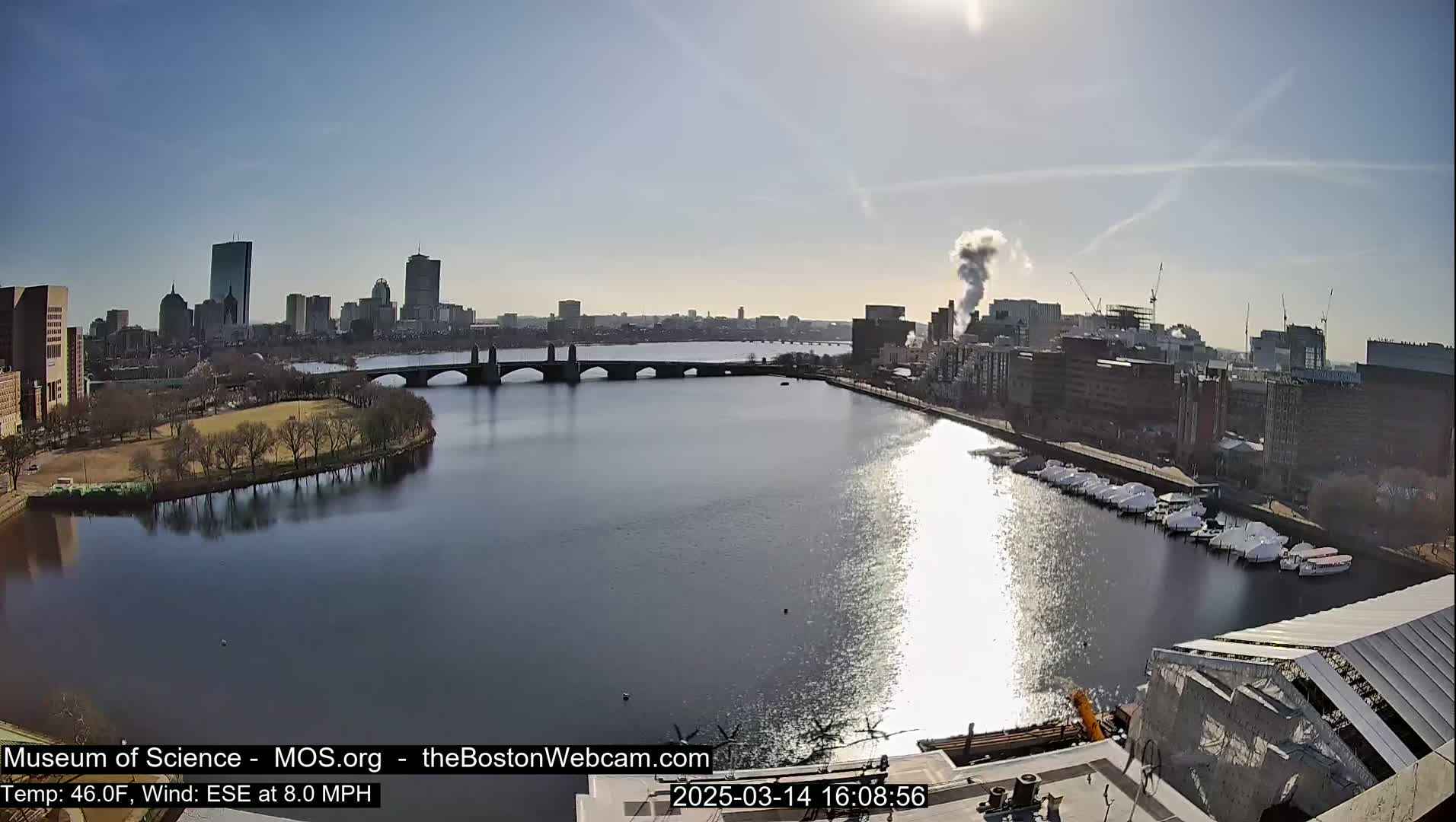 A sunny day shows a city skyline across a wide river, with a bridge and docked boats.