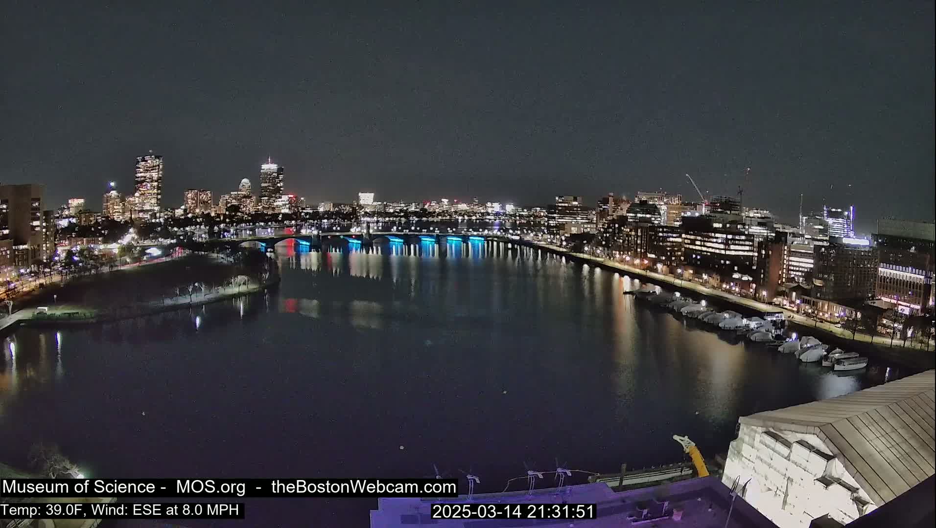 A nighttime view of a city skyline across a dark river, with a bridge illuminated in blue, under a clear sky.