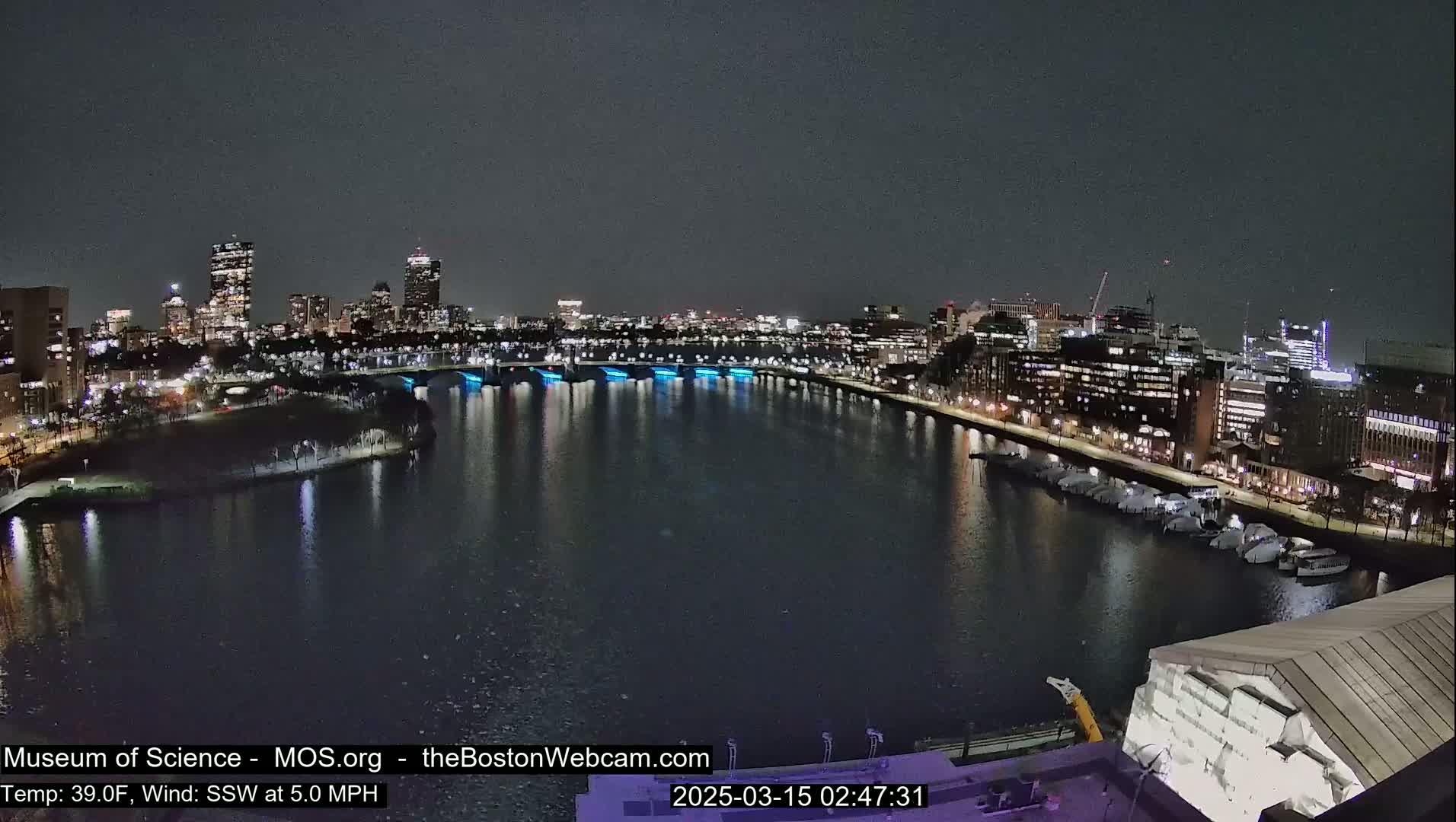 A nighttime view of a city skyline across a dark river, with boats docked along the shore and bridges illuminated with blue lights, under a clear, dark sky.